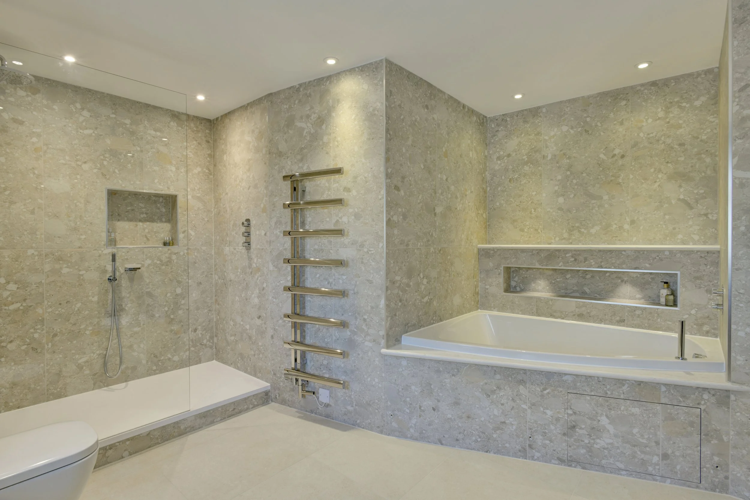 Modern bathroom with beige marble walls and floor, featuring a toilet, a bathtub, a glass-enclosed shower with a rainfall showerhead, and built-in shelves, with recessed ceiling lights.