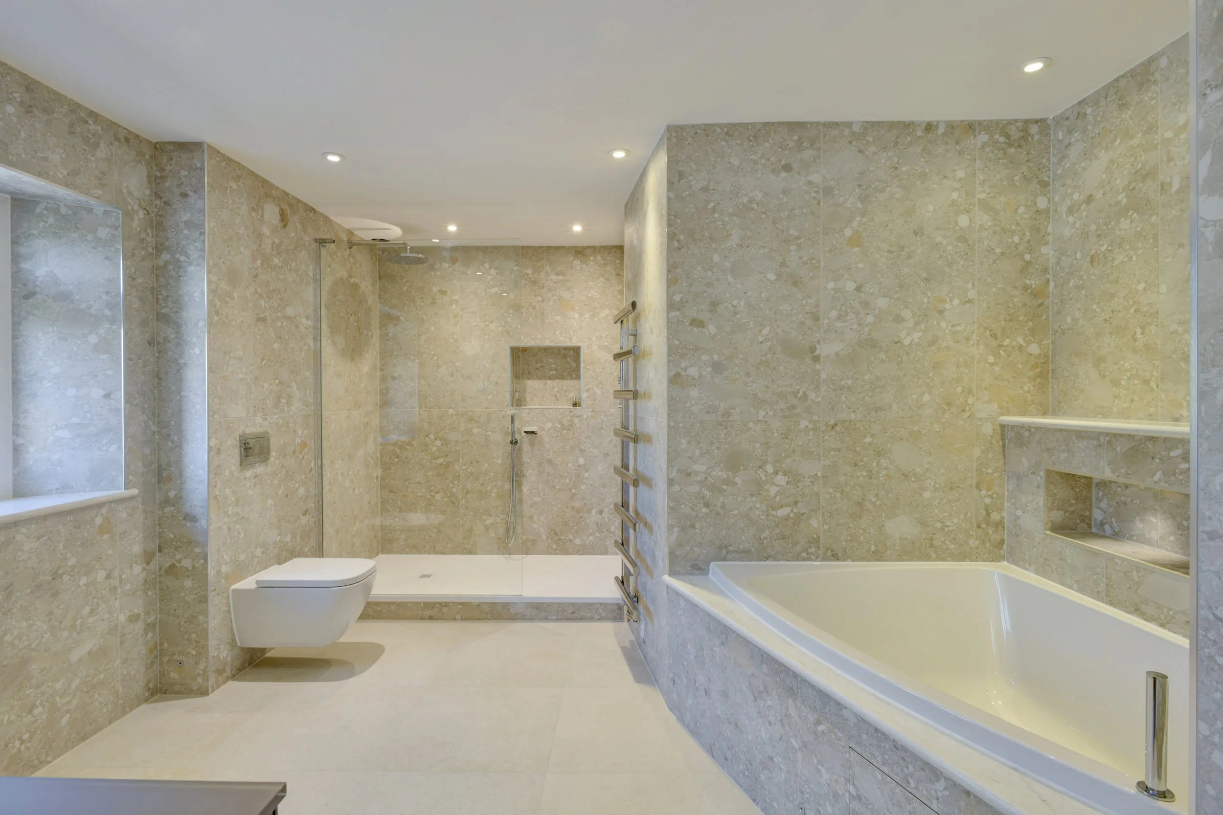 Modern bathroom with beige marble walls and floor, featuring a toilet, a bathtub, a glass-enclosed shower with a rainfall showerhead, and built-in shelves, with recessed ceiling lights.