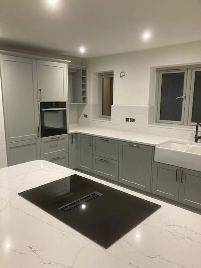 Modern kitchen with light gray cabinets, white marble countertops, built-in oven, black hob on island, white farmhouse sink, and two windows at the back.