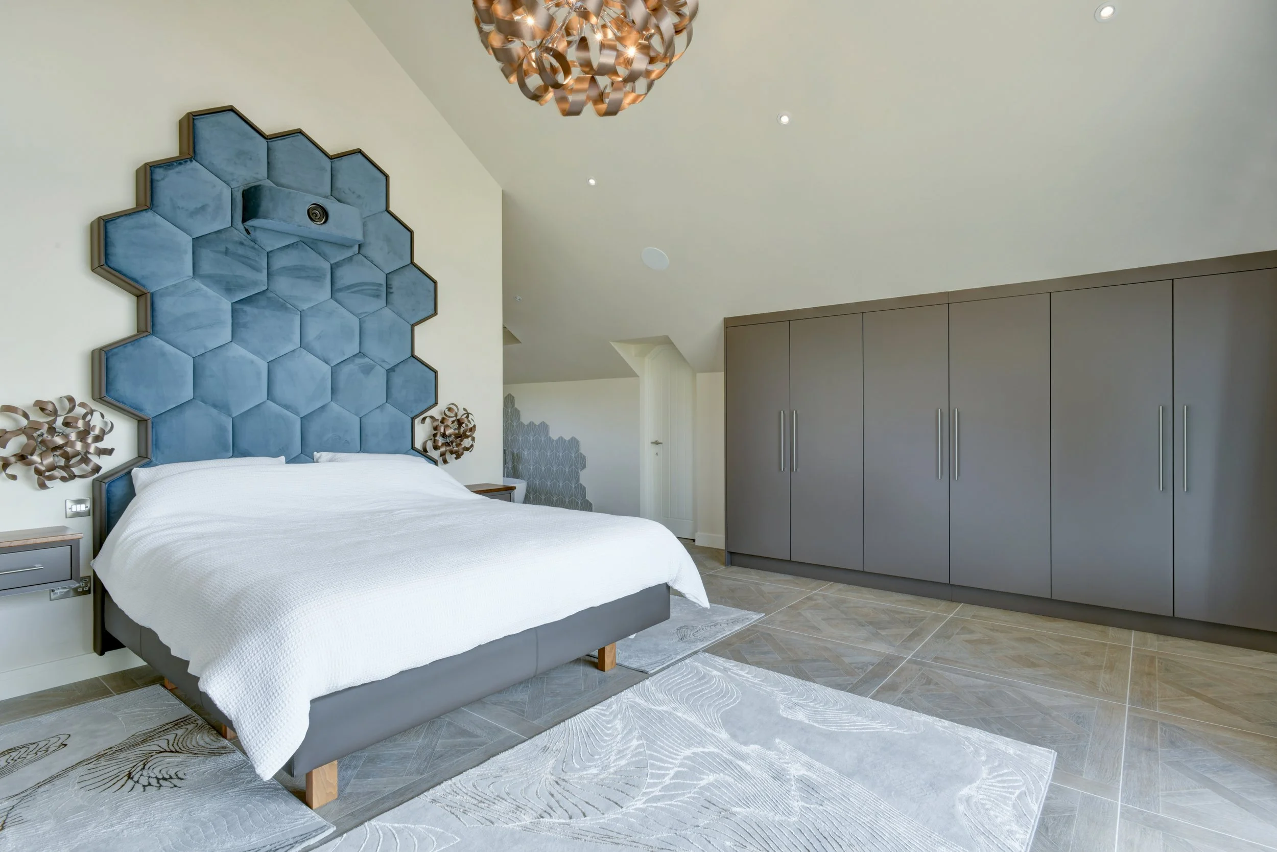 Modern bedroom with a bed, a large blue hexagonal headboard, and a built-in wardrobe, with a chandelier hanging from the ceiling and decorative wall art.