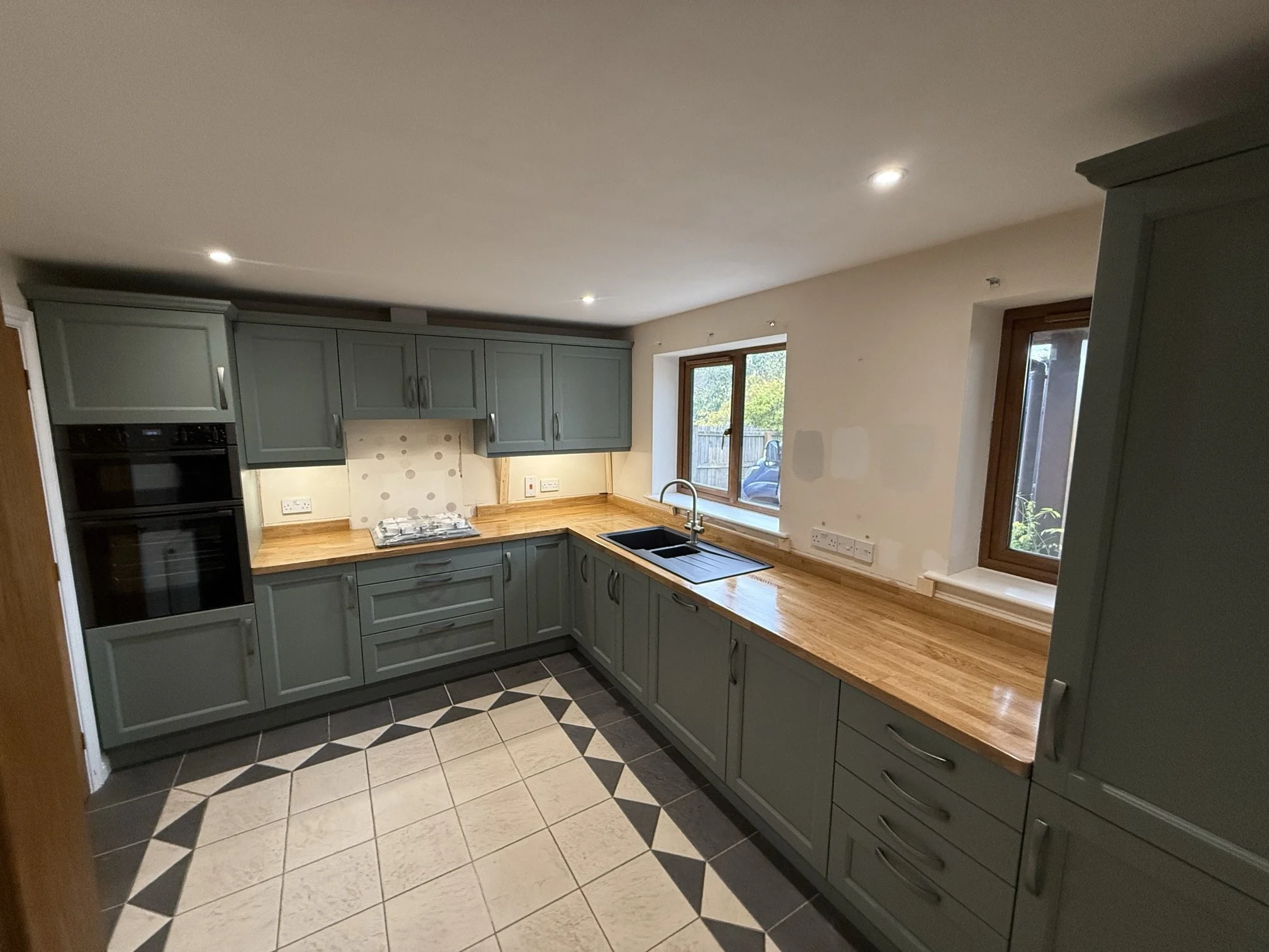 Modern kitchen with green cabinets, wooden countertop, black sink, and a window overlooking the outside. The floor has a patterned tile design.