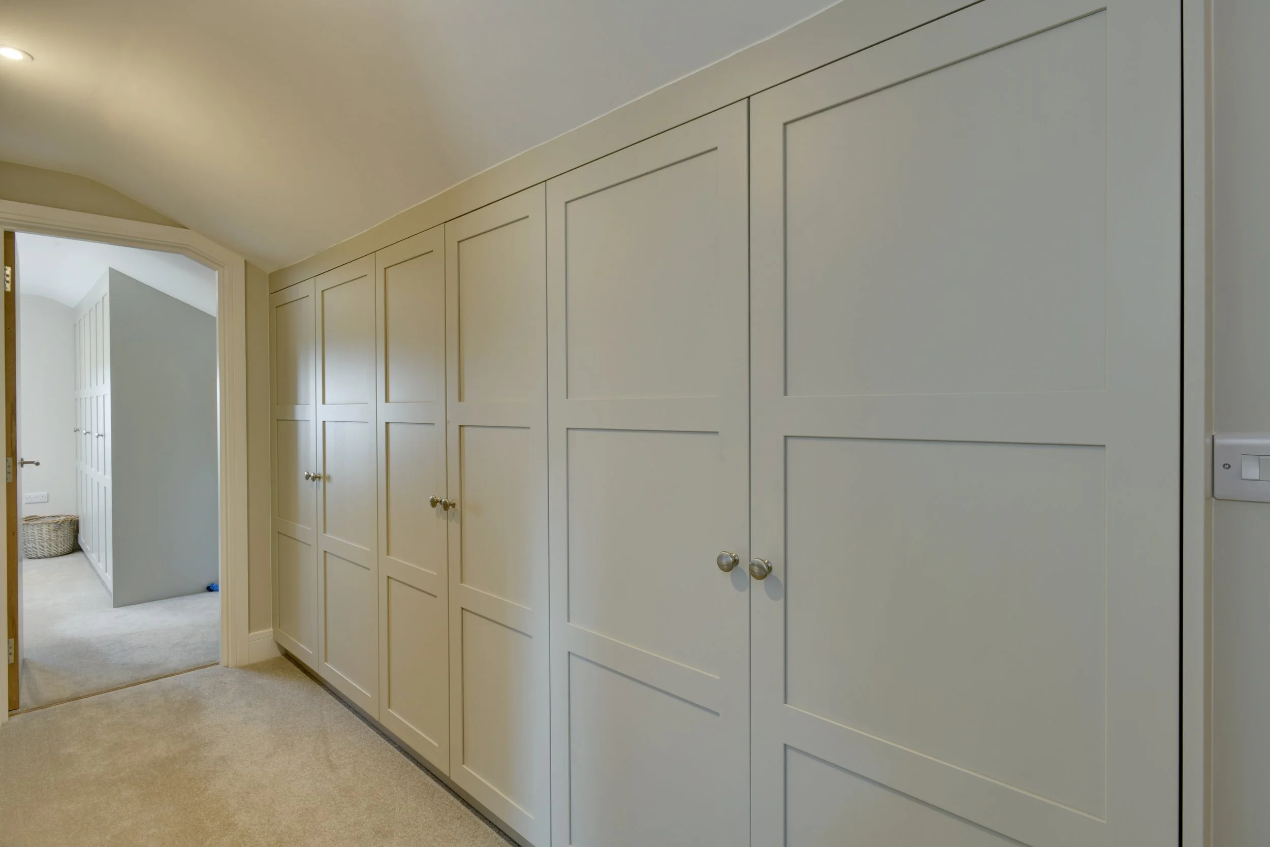 Off-White built-in wardrobes with silver round knobs in a narrow hallway with beige carpeting and light-colored walls.
