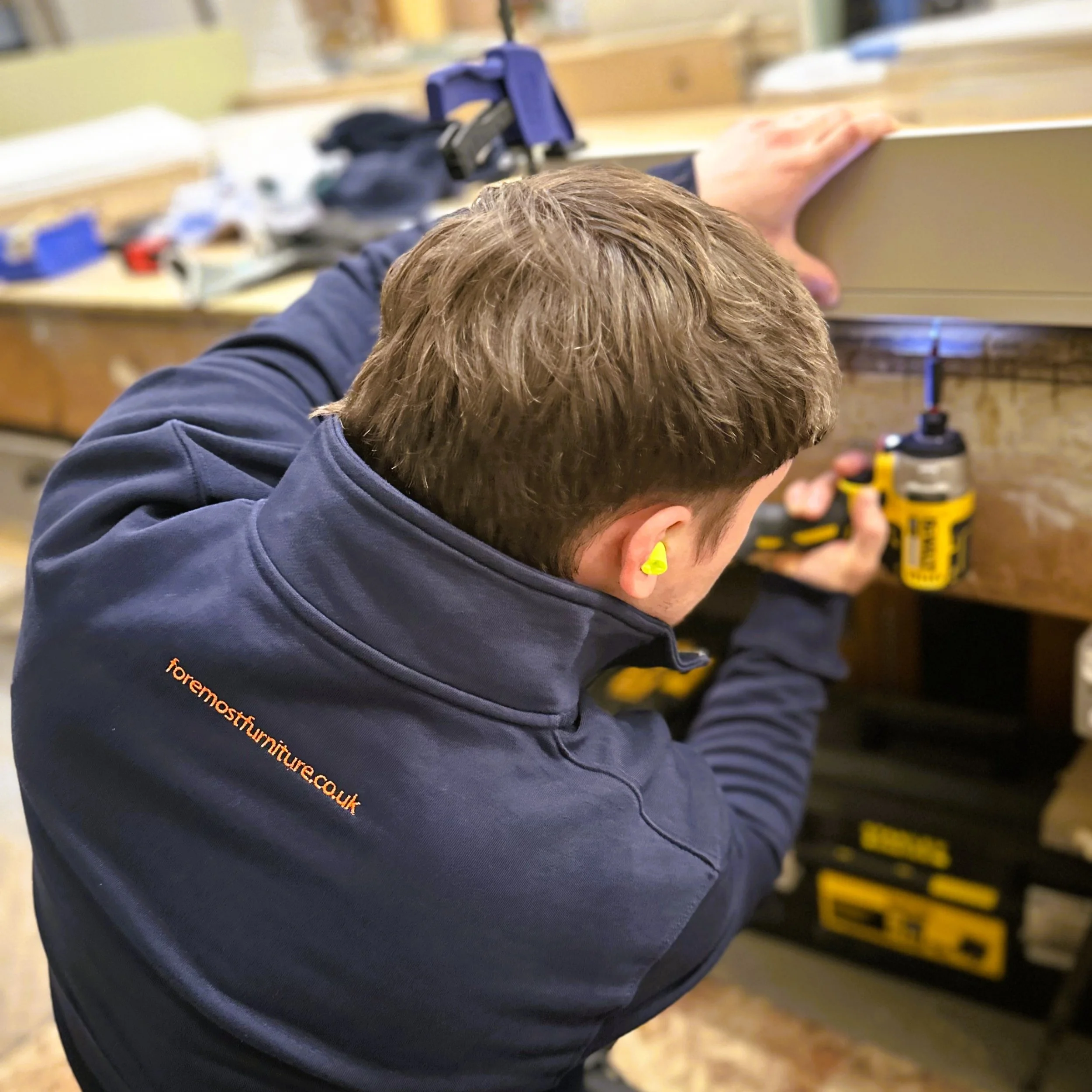 A person working on a project in a workshop, using a power tool on a piece of wood or similar material, with various tools and equipment in the background.