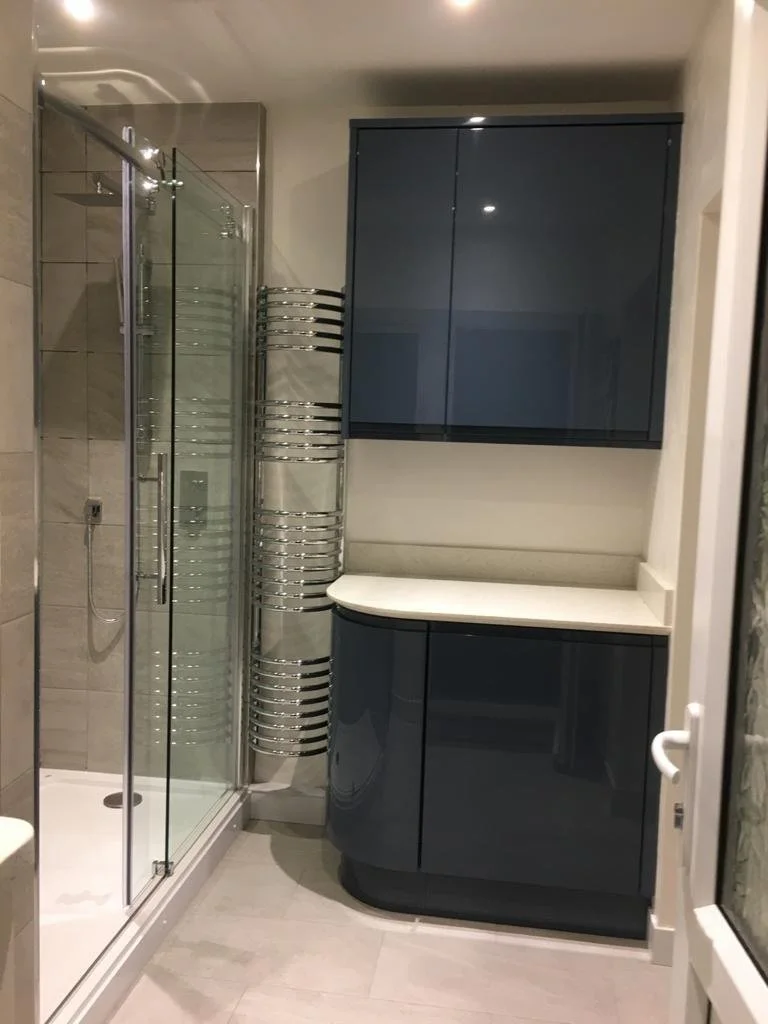 A modern bathroom with a glass shower enclosure, a built-in shower head, a curved dark blue cabinet with a white countertop, and a vertically mounted heated towel rack.