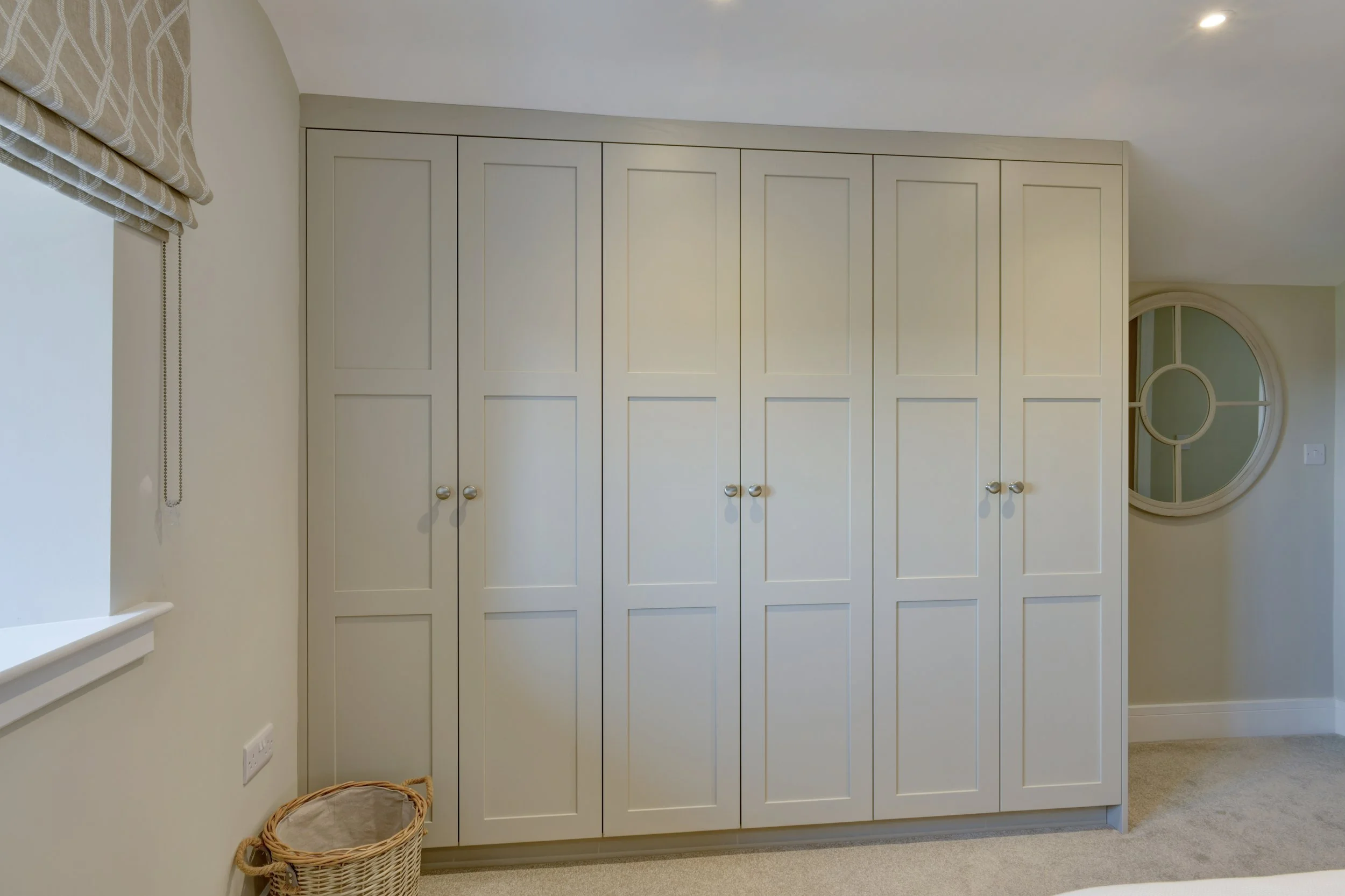 A large white built-in wardrobe with framed panel doors and round metallic knobs. There is a beige basket on the floor near the wall and a window with a beige patterned Roman shade on the left.