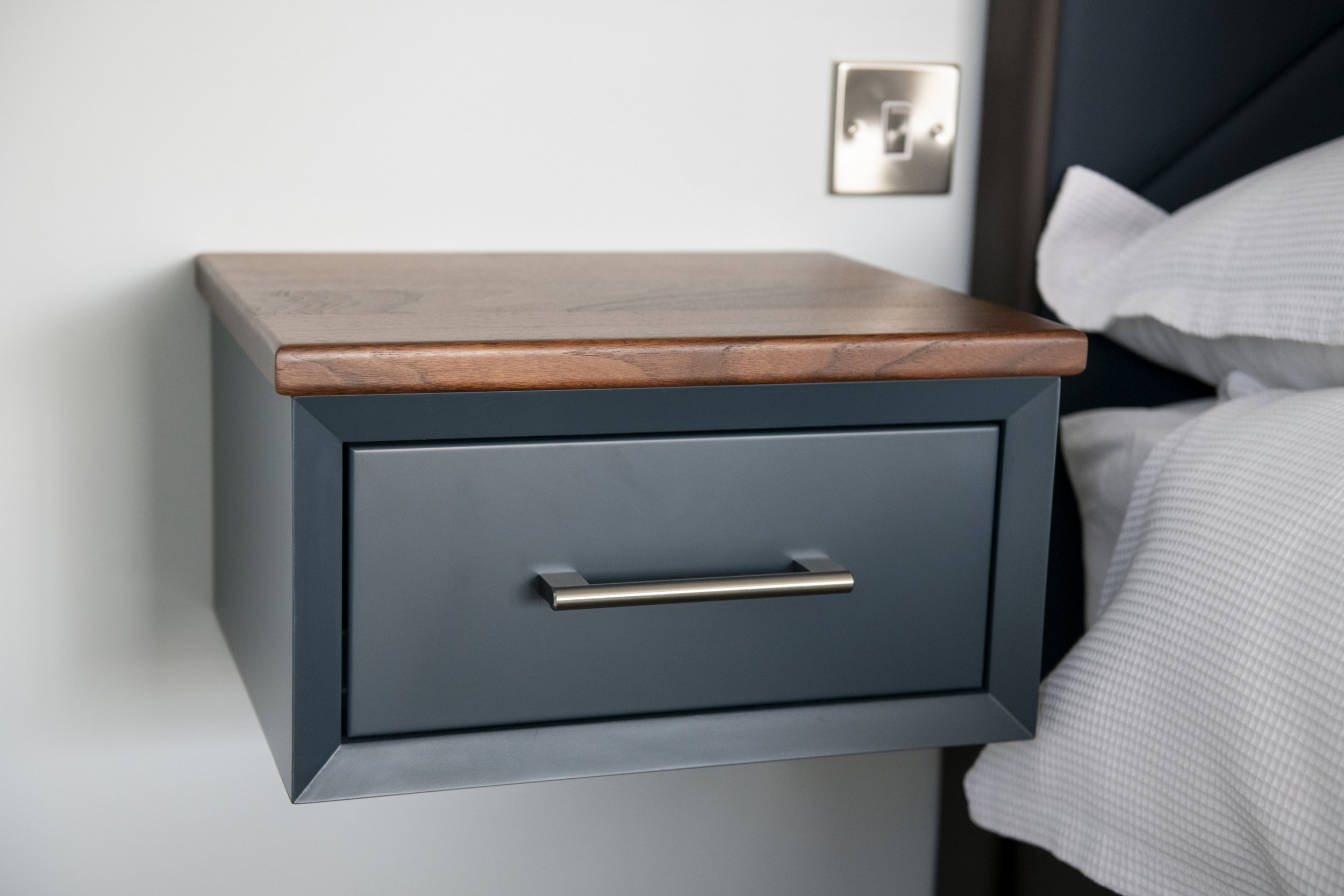 A floating bedside tablewith a wooden top and dark blue drawer, next to a bed.