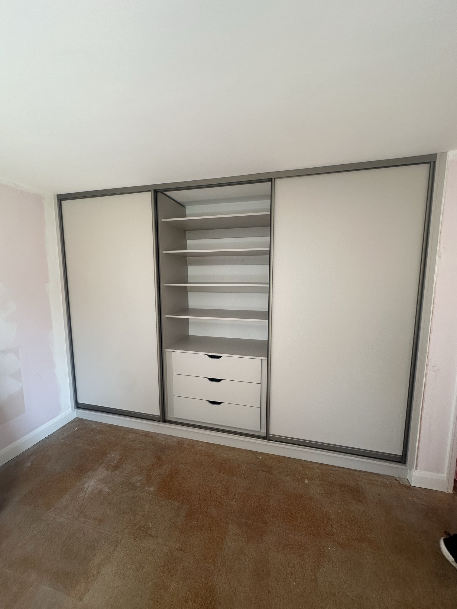 A white built-in wardrobe with sliding doors, one with shelves and drawers inside, situated against a wall in a room with brown flooring.