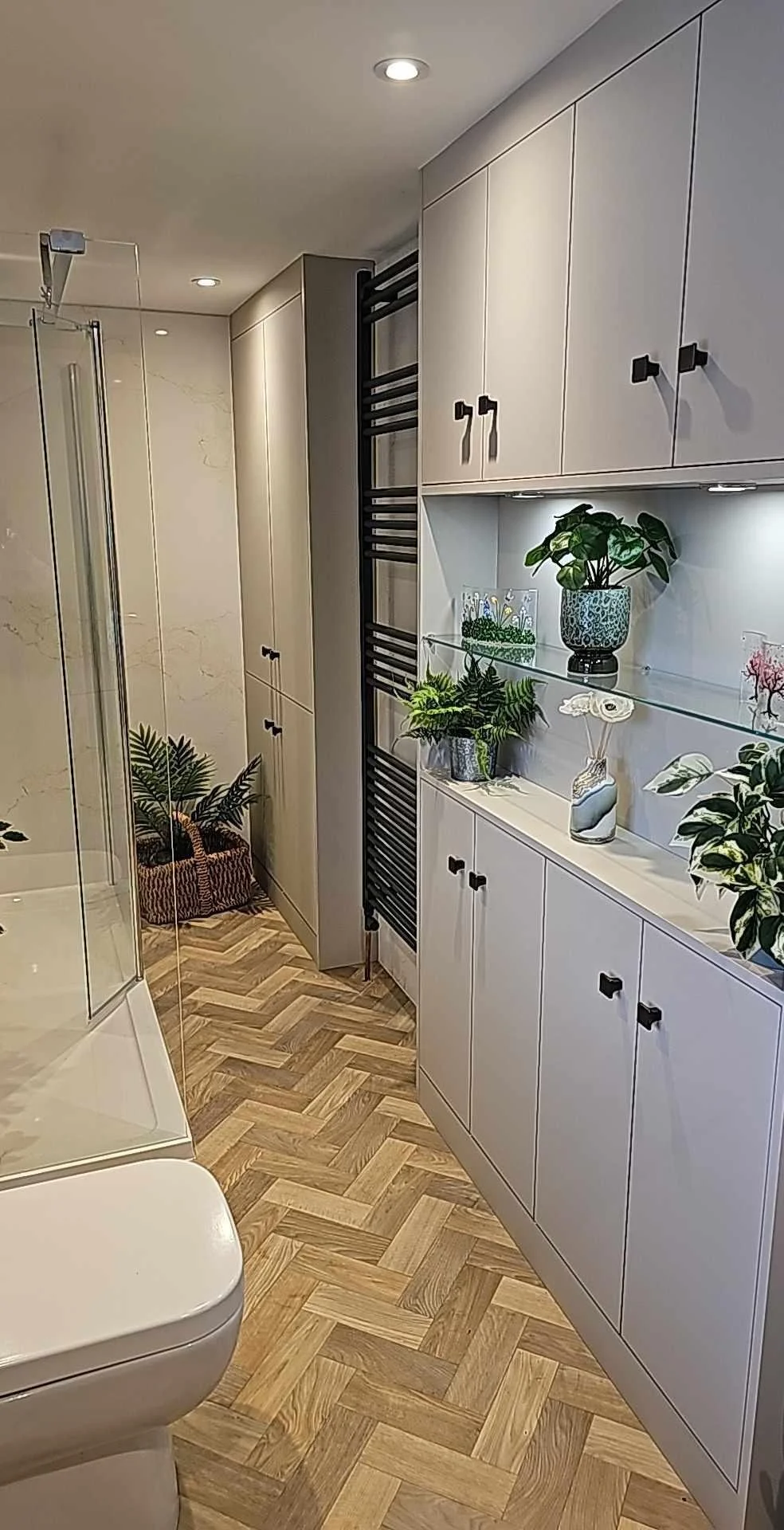 Modern bathroom with white cabinets, glass display shelf containing plants and decorative items, black radiator, wooden herringbone flooring, shower enclosure, and a white toilet.