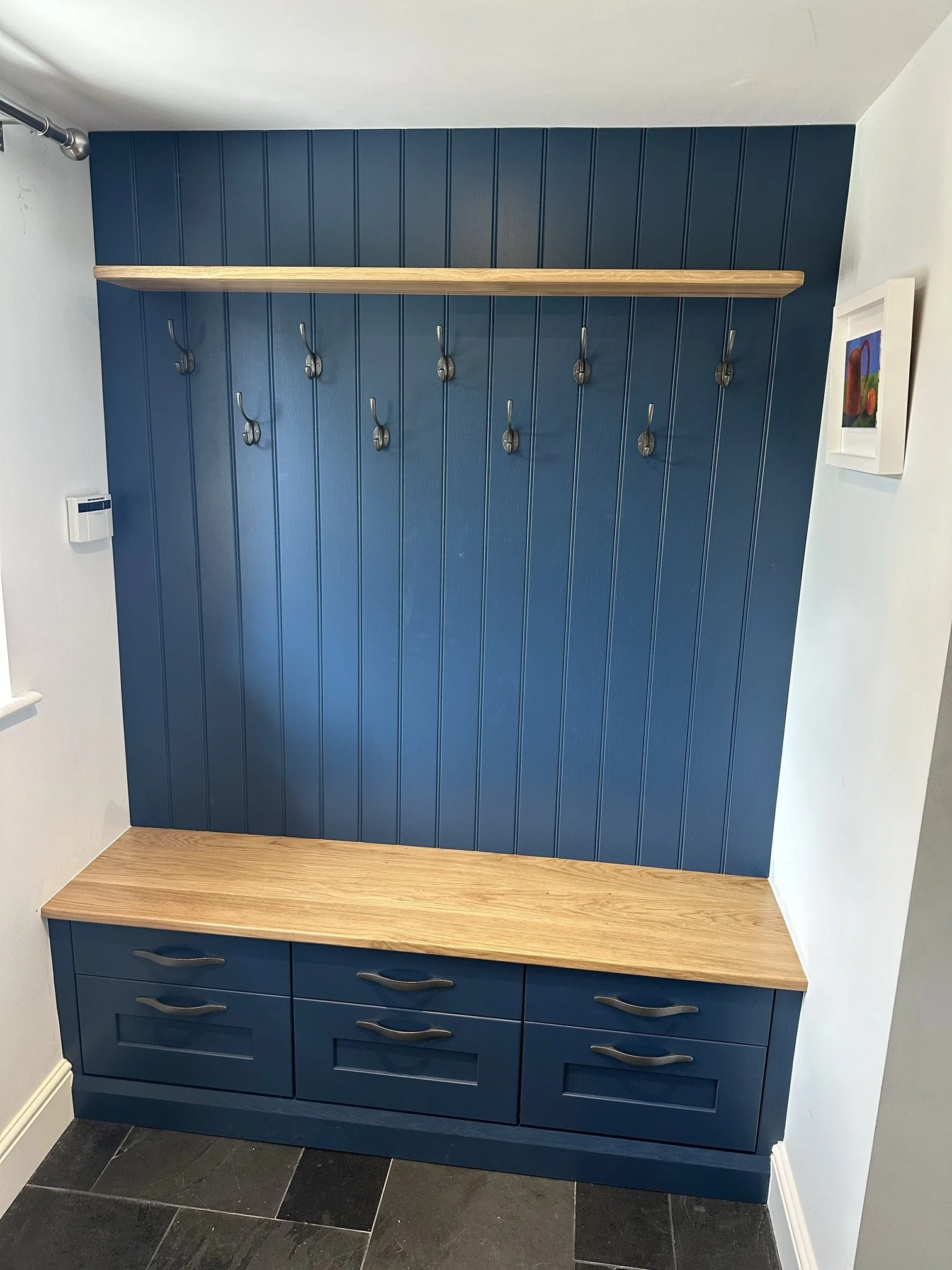 A blue boot room, including a wooden shelf with coat hooks underneath. There is a shoe cabinet with drawers and a wooden bench beneath.