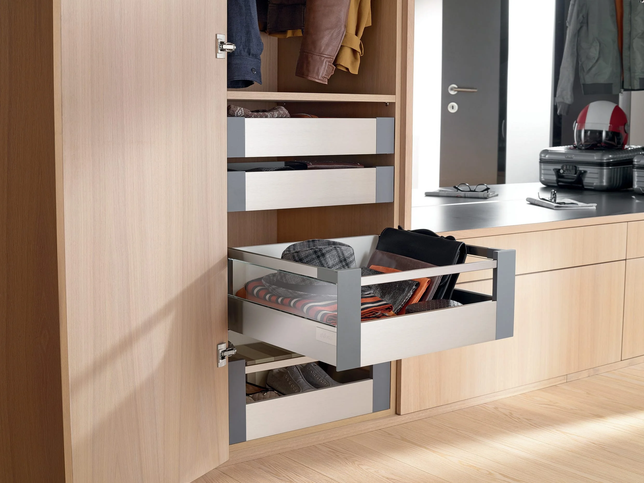 Blum open wooden wardrobe with pull-out drawers and shelves containing folded clothes, a hat, and shoes, located in a modern room with a desk and luggage.