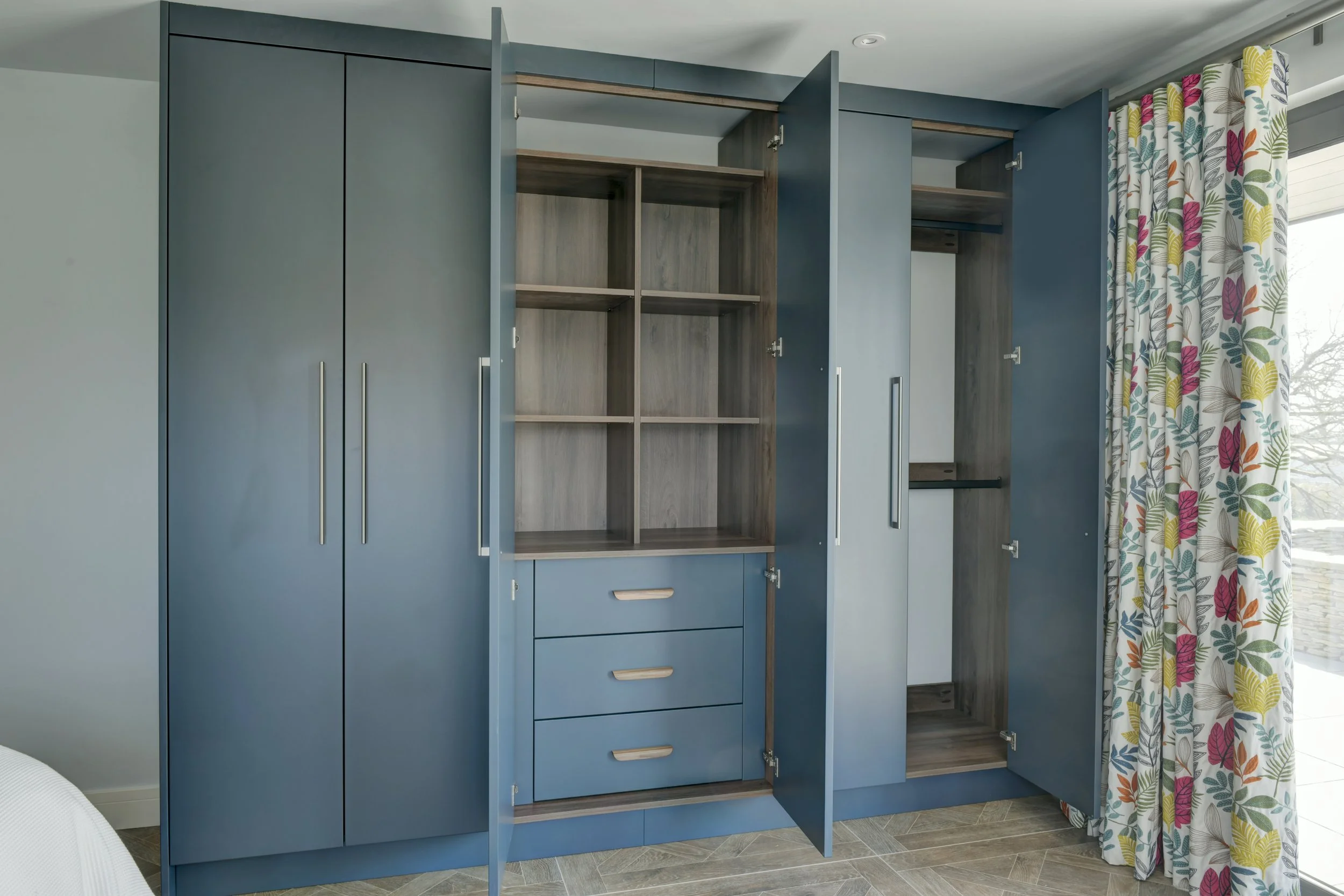Blue wardrobe with open doors revealing internal shelves. Curtain with colorful leaf pattern near a large window.