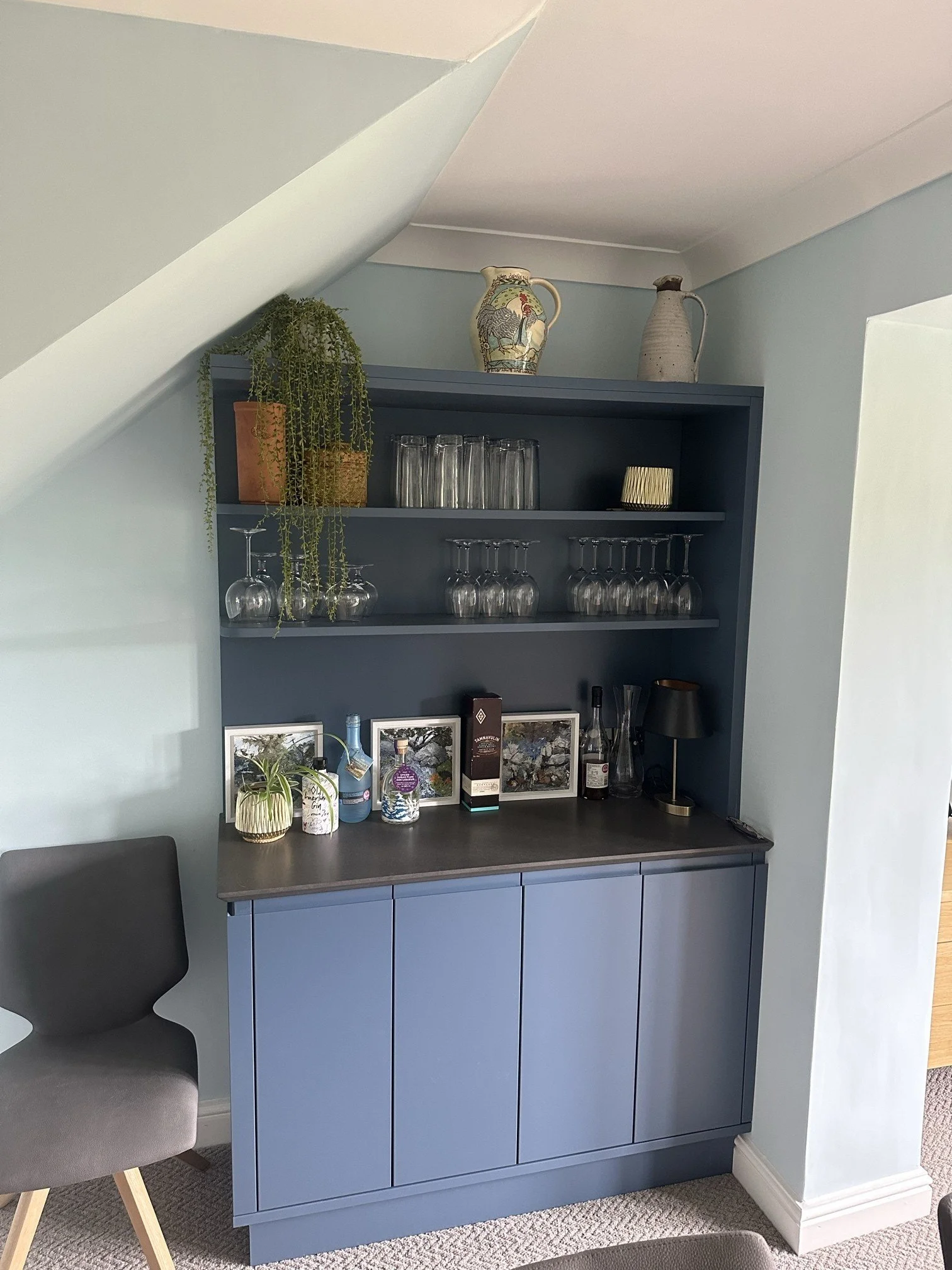 A blue bar cabinet with artwork, glassware, and decorative items on top, adjacent to a dark grey chair with wooden legs.
