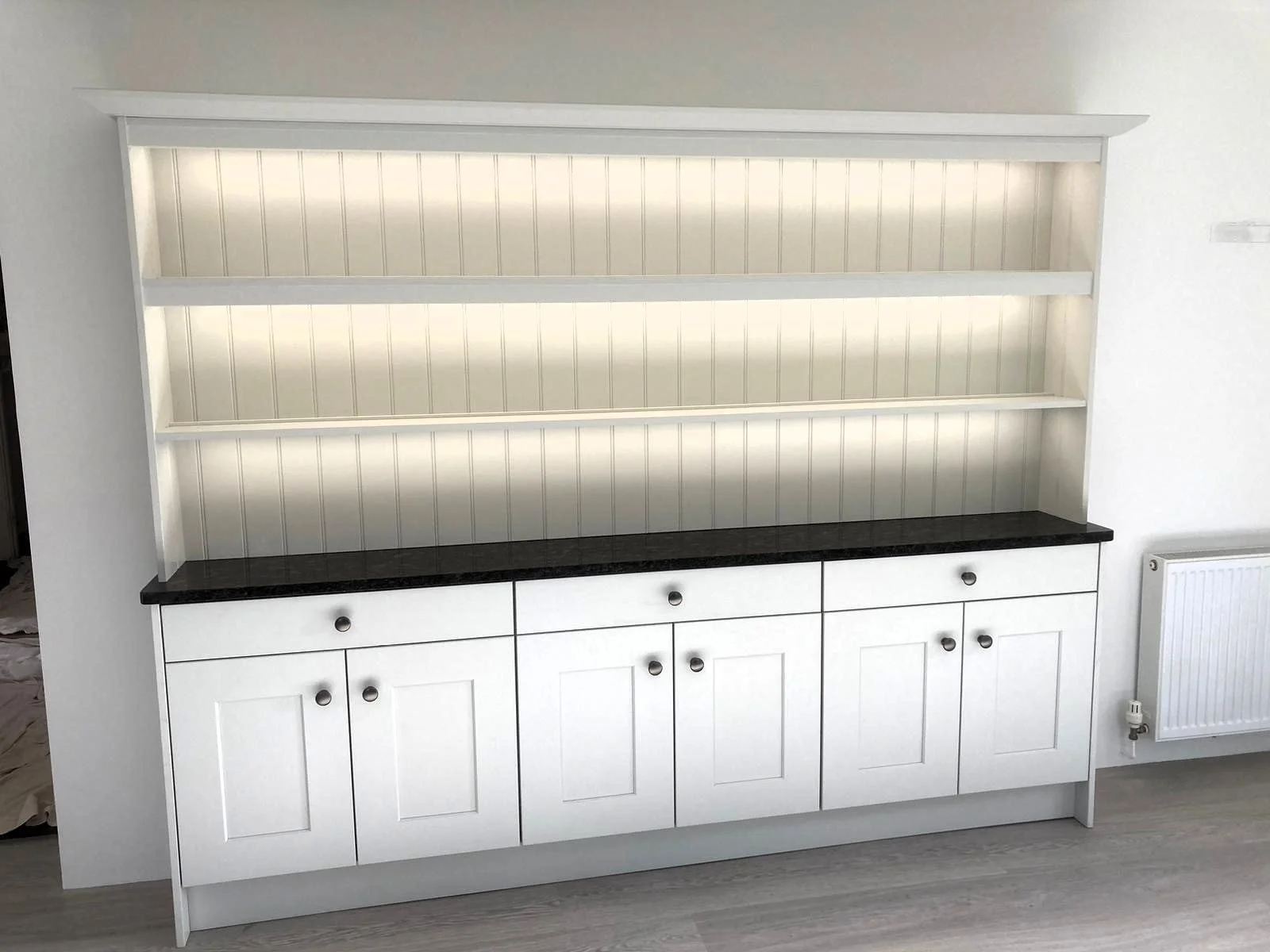 A white unit with three open shelves, black countertop, and storage cabinets below, positioned against a white wall with a radiator nearby.