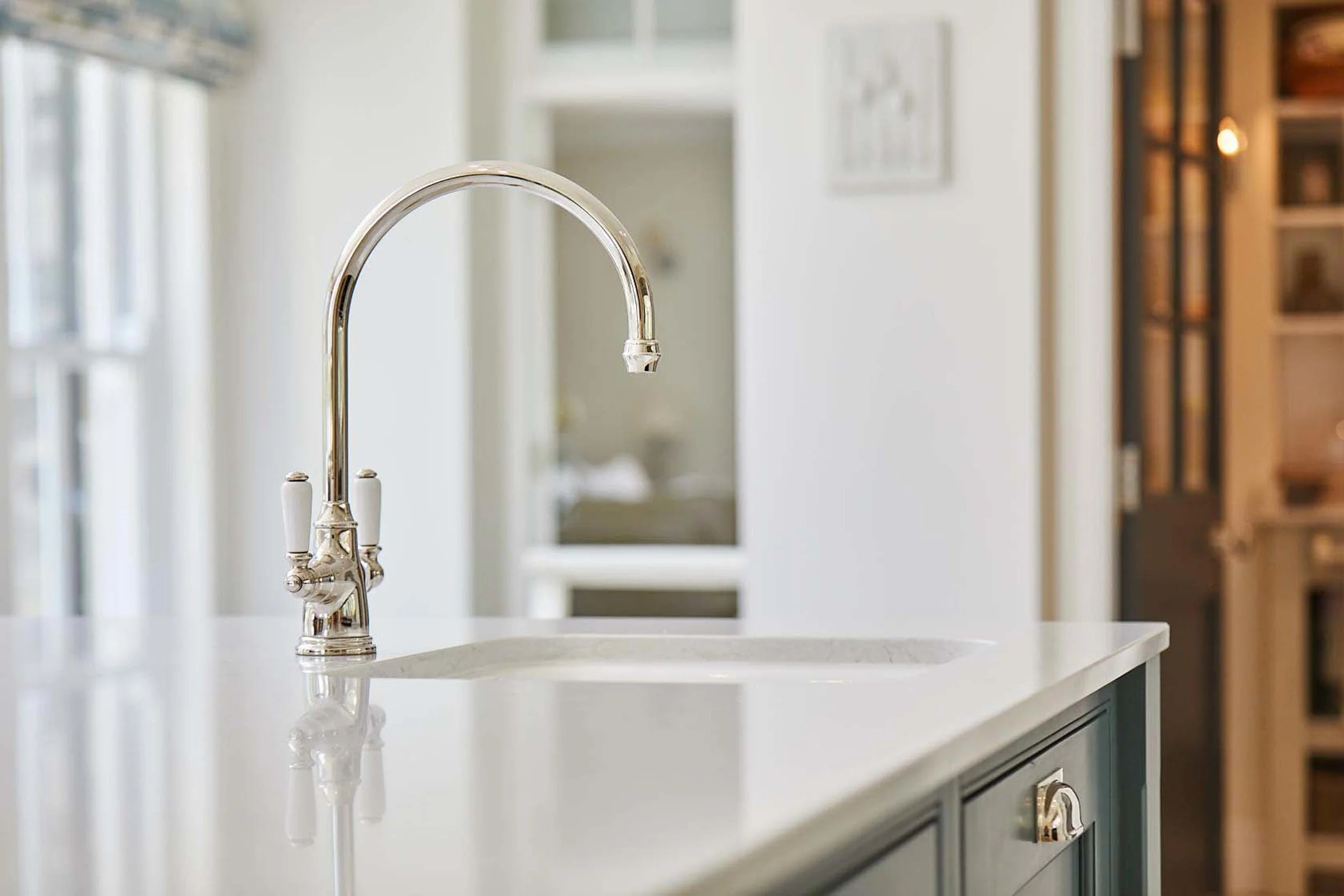 Close-up of a kitchen island with a Perrin & Roww polished chrome faucet and white sink. With sunlight streaming through the windows and blurred background of kitchen cabinetry and decor.