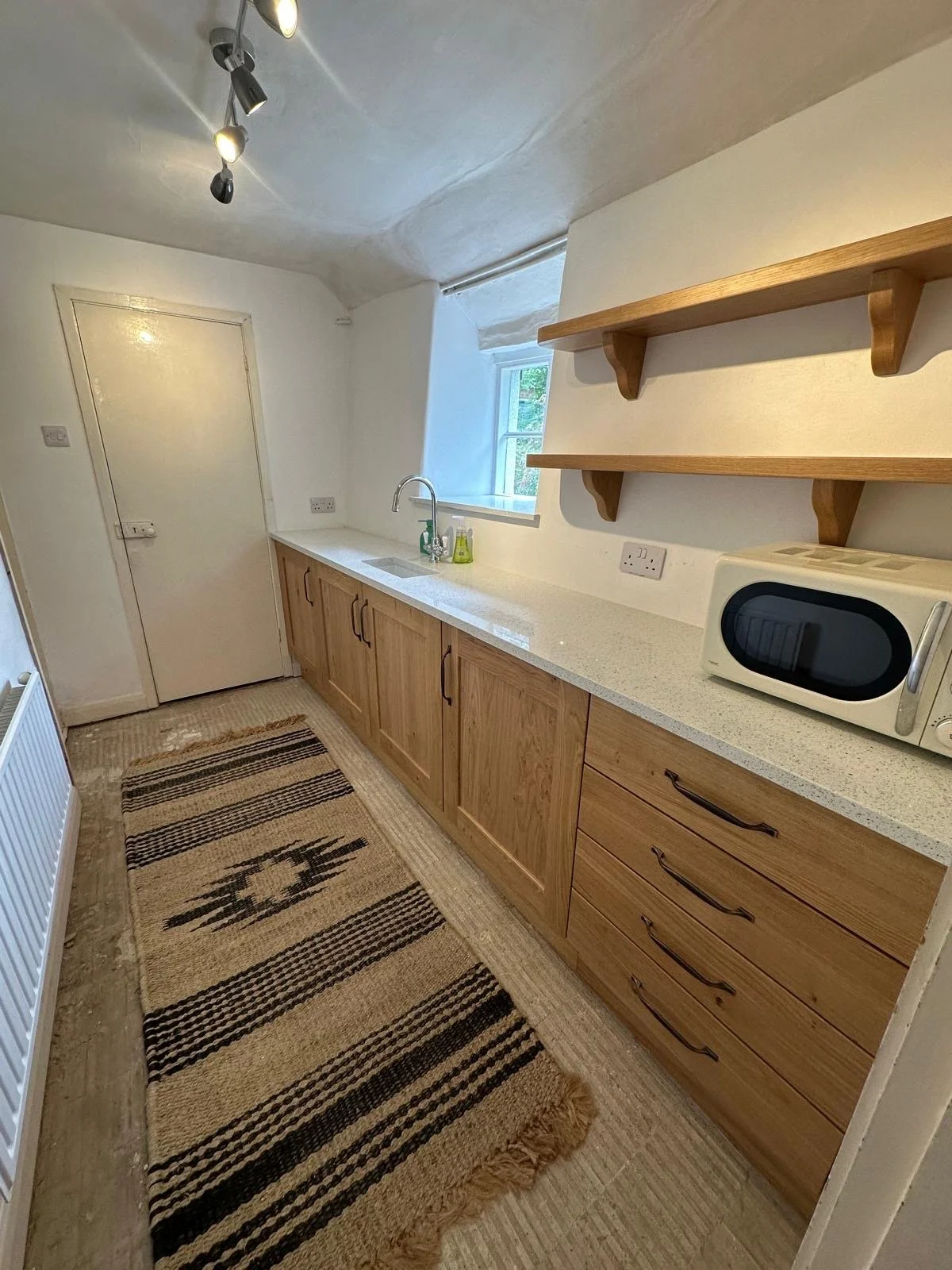 A utility room with wooden cabinets, a white countertop, and open wooden shelves. There is a microwave on the counter, a window above the sink, and a striped rug on the floor.