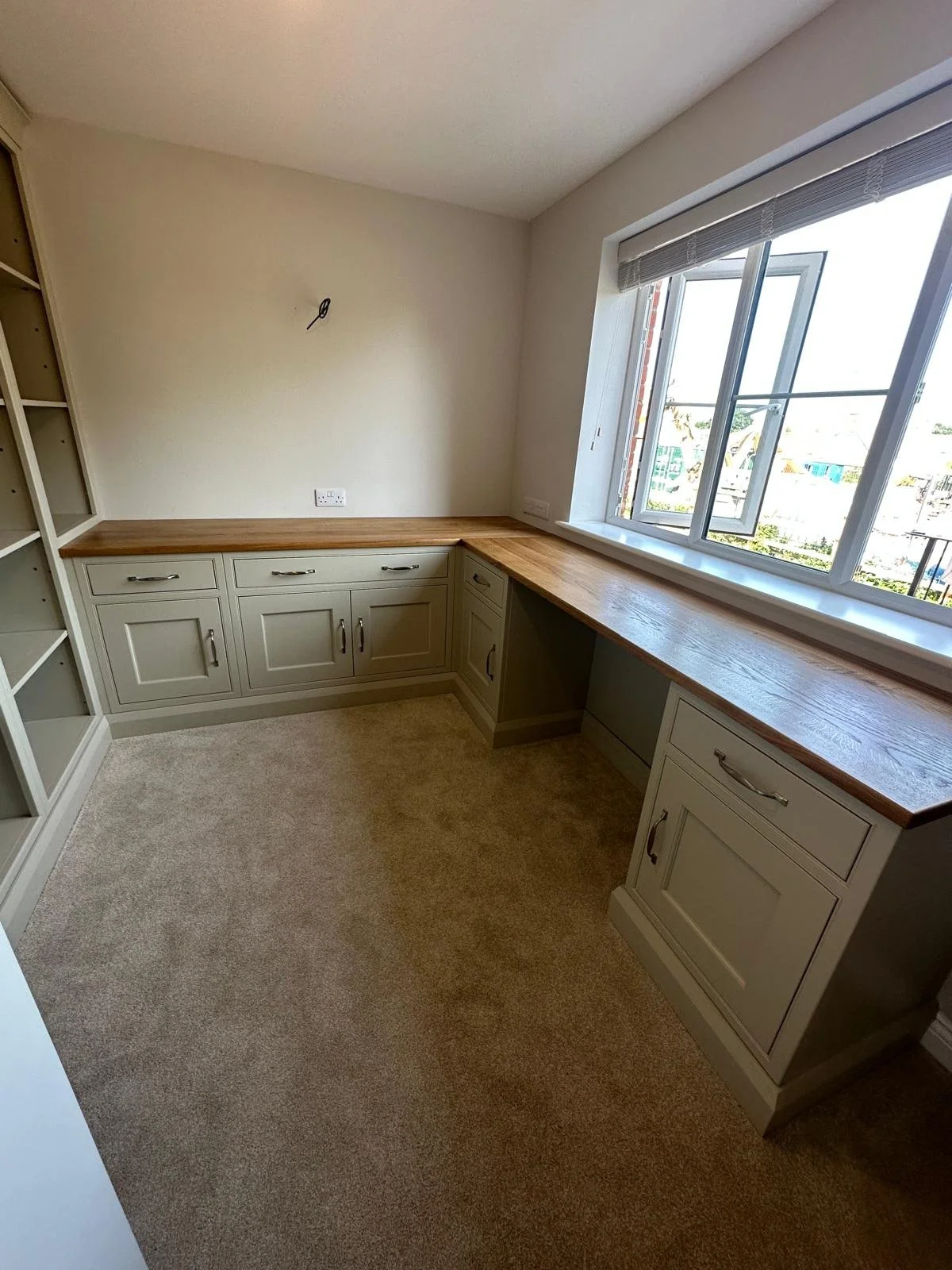 Office with built-in desk and shelves, a wooden countertop, grey cabinets, a large window with blinds, and beige carpet flooring.
