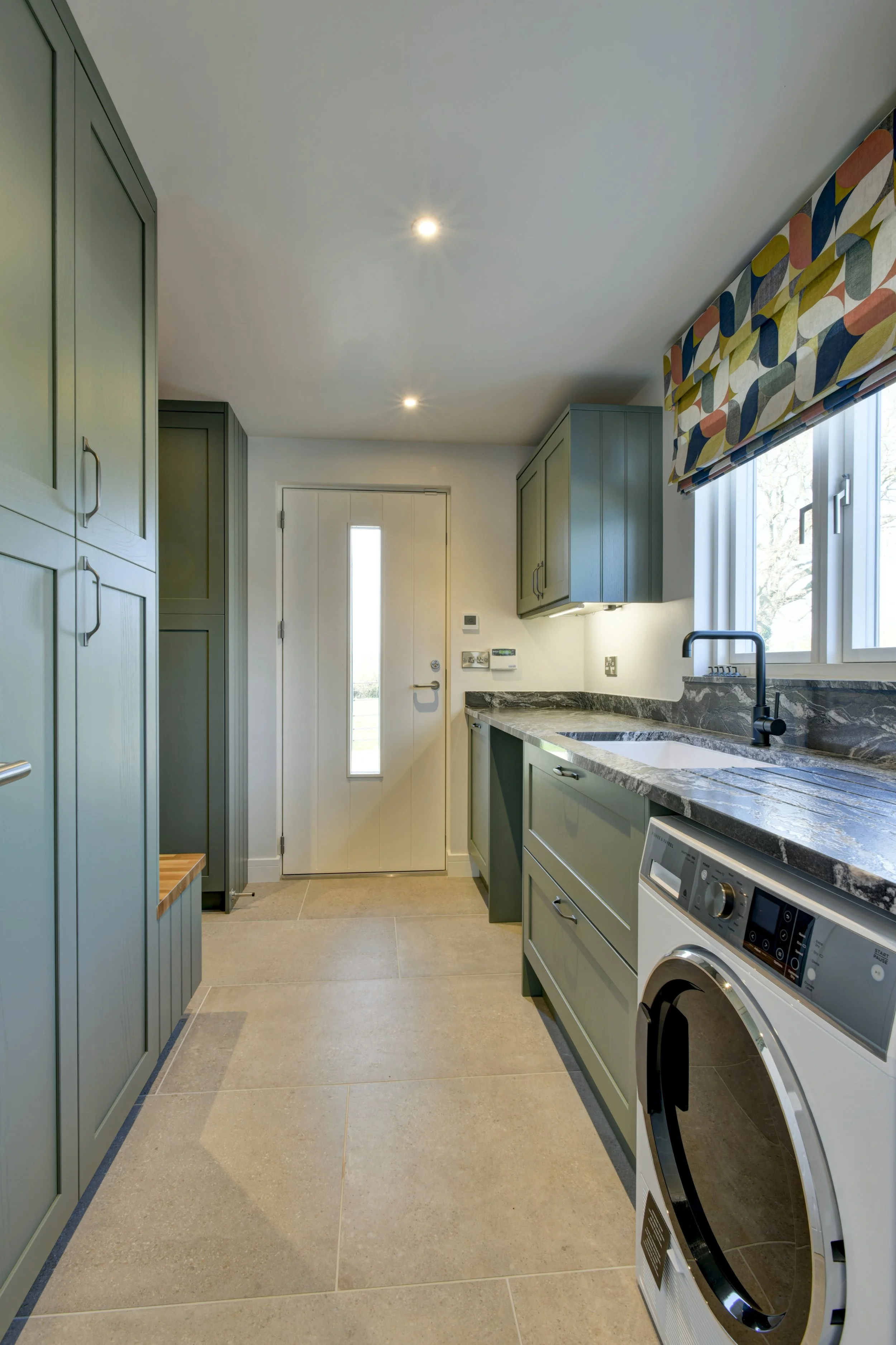 A modern laundry room with green cabinets, a marble countertop, a washing machine, a window with a colorful patterned Roman shade, a door with a narrow window, and recessed ceiling lights.