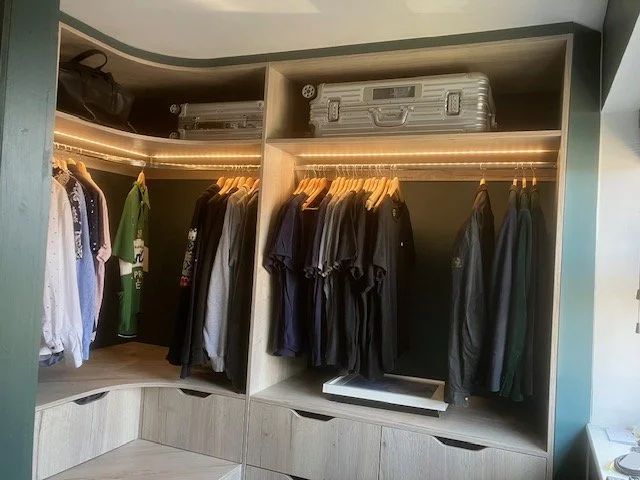 A closet with hanging clothes including shirts and jackets, with suitcases stored on a shelf above.