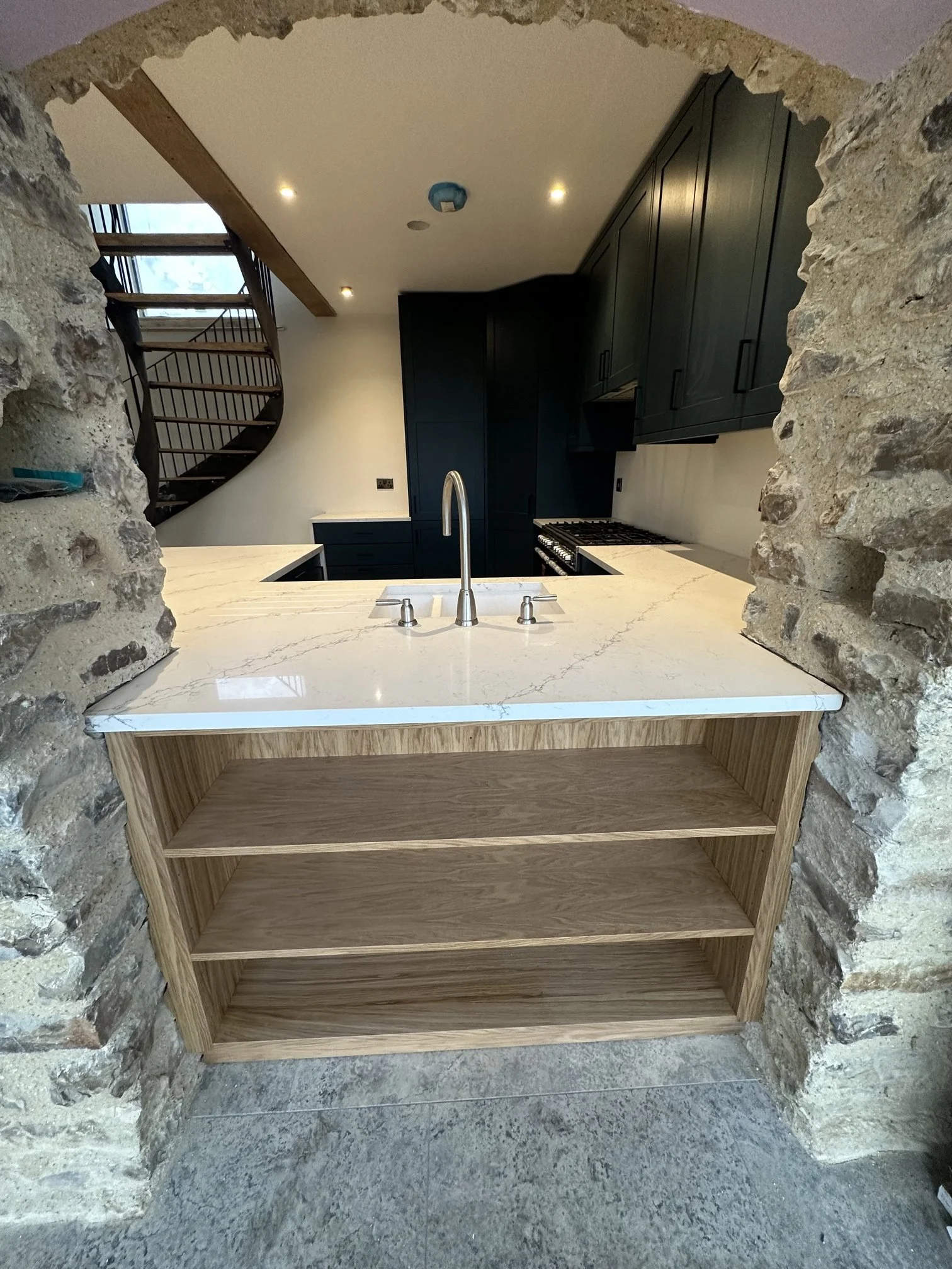 View of a kitchen through an stone wall opening, featuring a marble countertop with a modern faucet, dark cabinets, and a wooden staircase visible in the background.