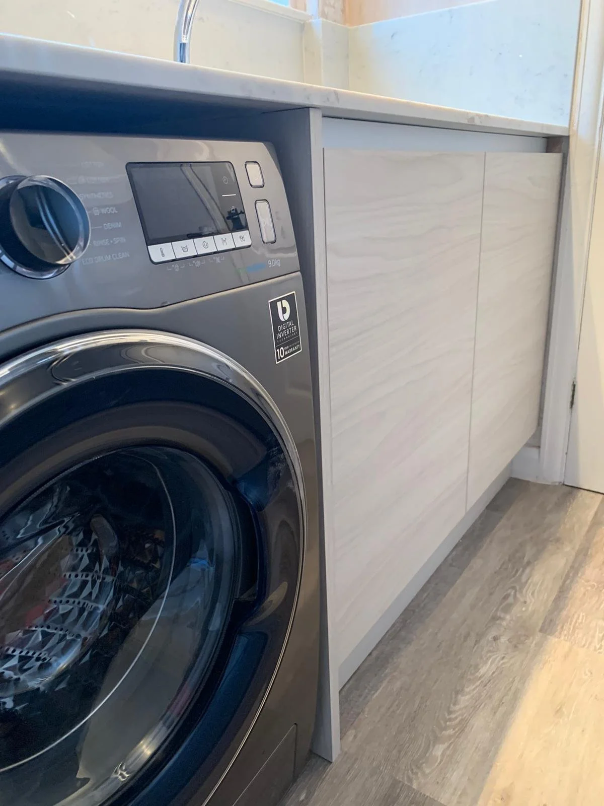A front-loading washing machine with a digital display, placed next to a white cabinet with a marble countertop in a laundry room.