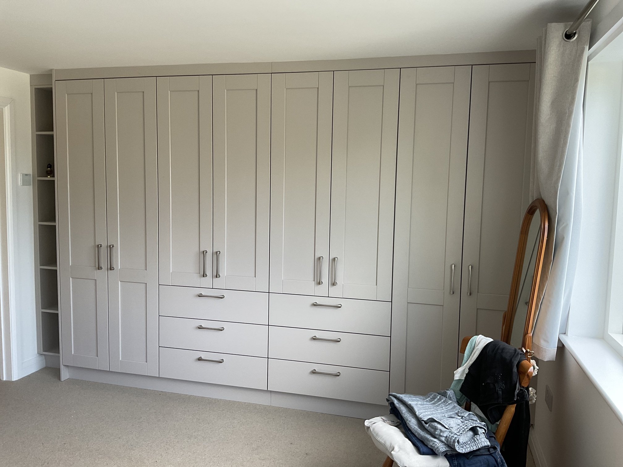 A large wardrobe with multiple doors and drawers, situated in a room with beige carpet, next to a window with a curtain, and a chair holding clothing and a mirror nearby.