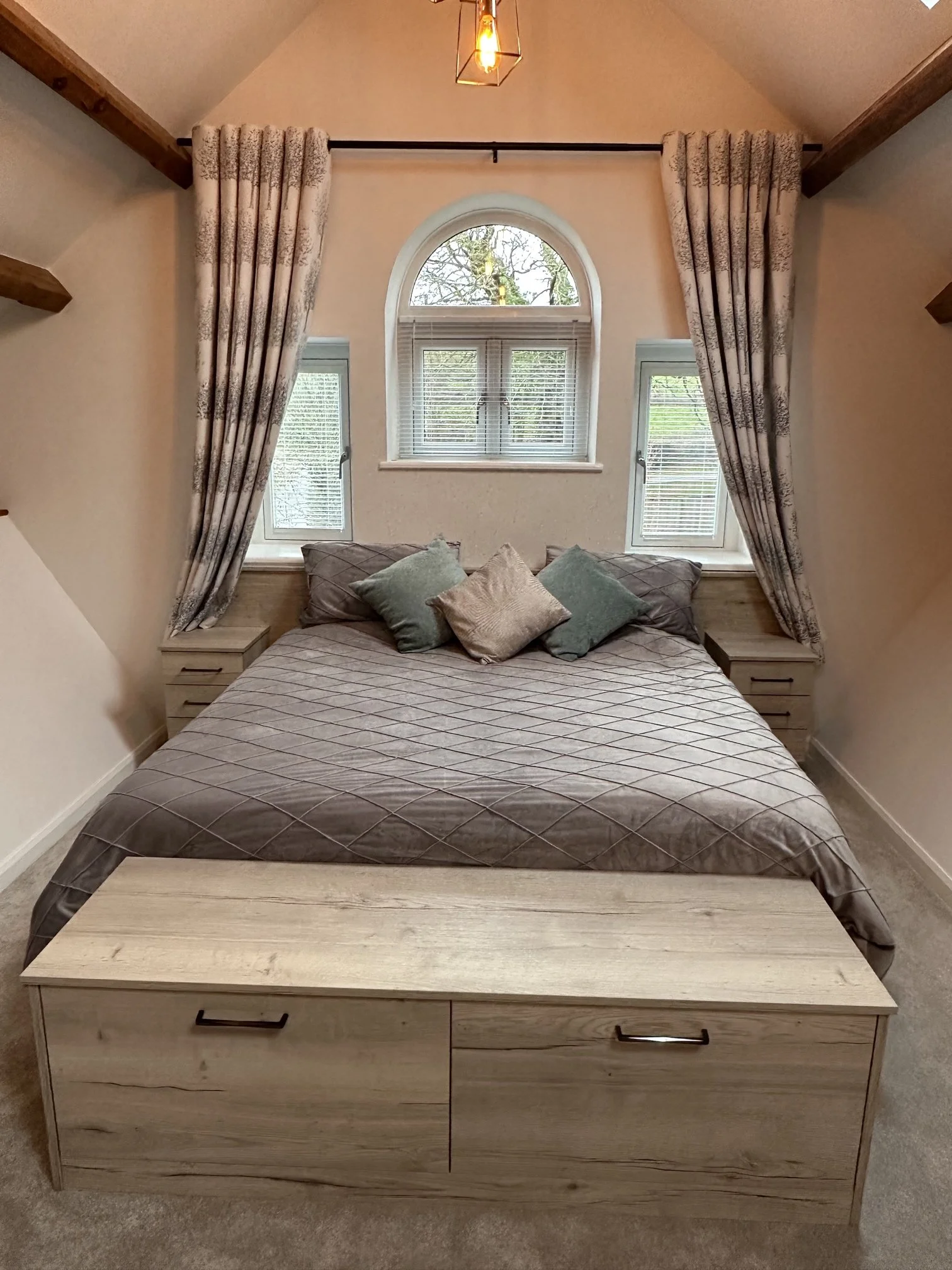 A bedroom with a large bed covered in a brown quilted bedspread and multiple pillows, wood nightstands on each side, a window with an arched top and blinds, curtains framing the window, a wooden storage bench at the foot of the bed.