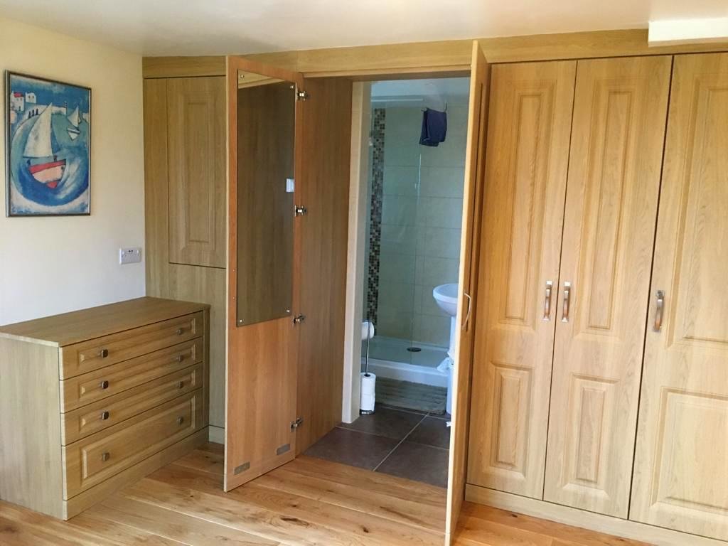 Built in wardrobe including a chest of drawers and large cupboards, an open door revealing a bathroom with a shower and sink, and a painting on the wall.