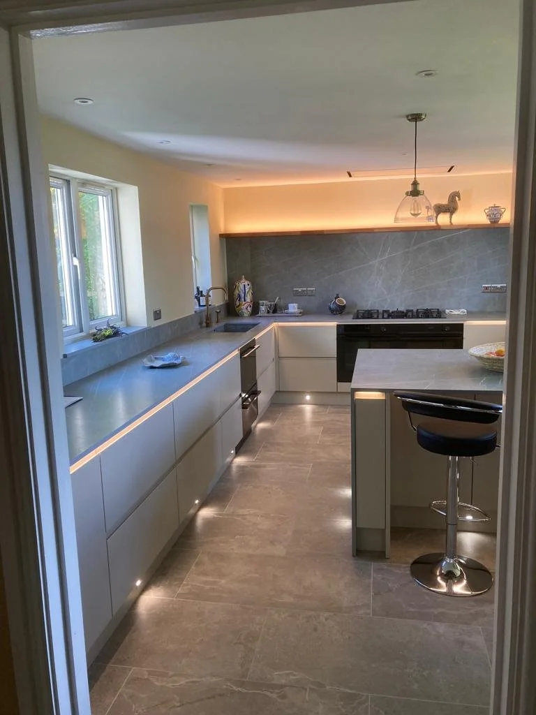 Modern kitchen with white cabinets and grey countertops, illuminated by under-cabinet and ceiling lighting.