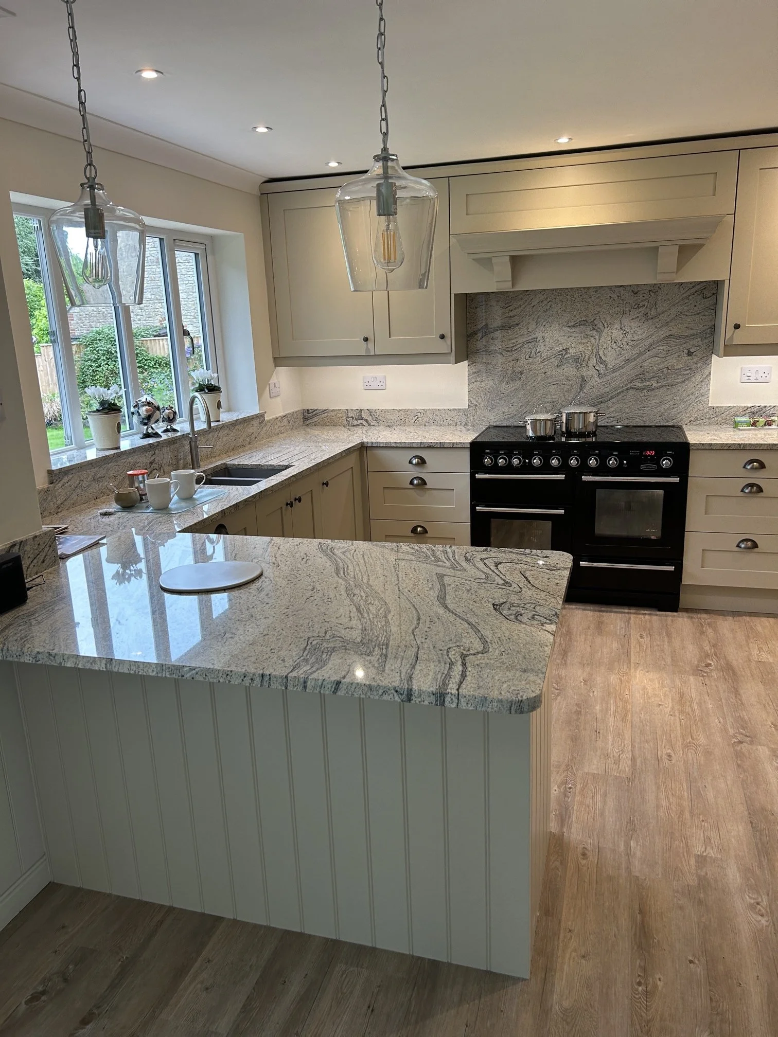 A kitchen with beige cabinetry, a black range cooker, granite countertops, and a window with a view of greenery outside. Decor includes small potted plants and decorative items on the windowsill.