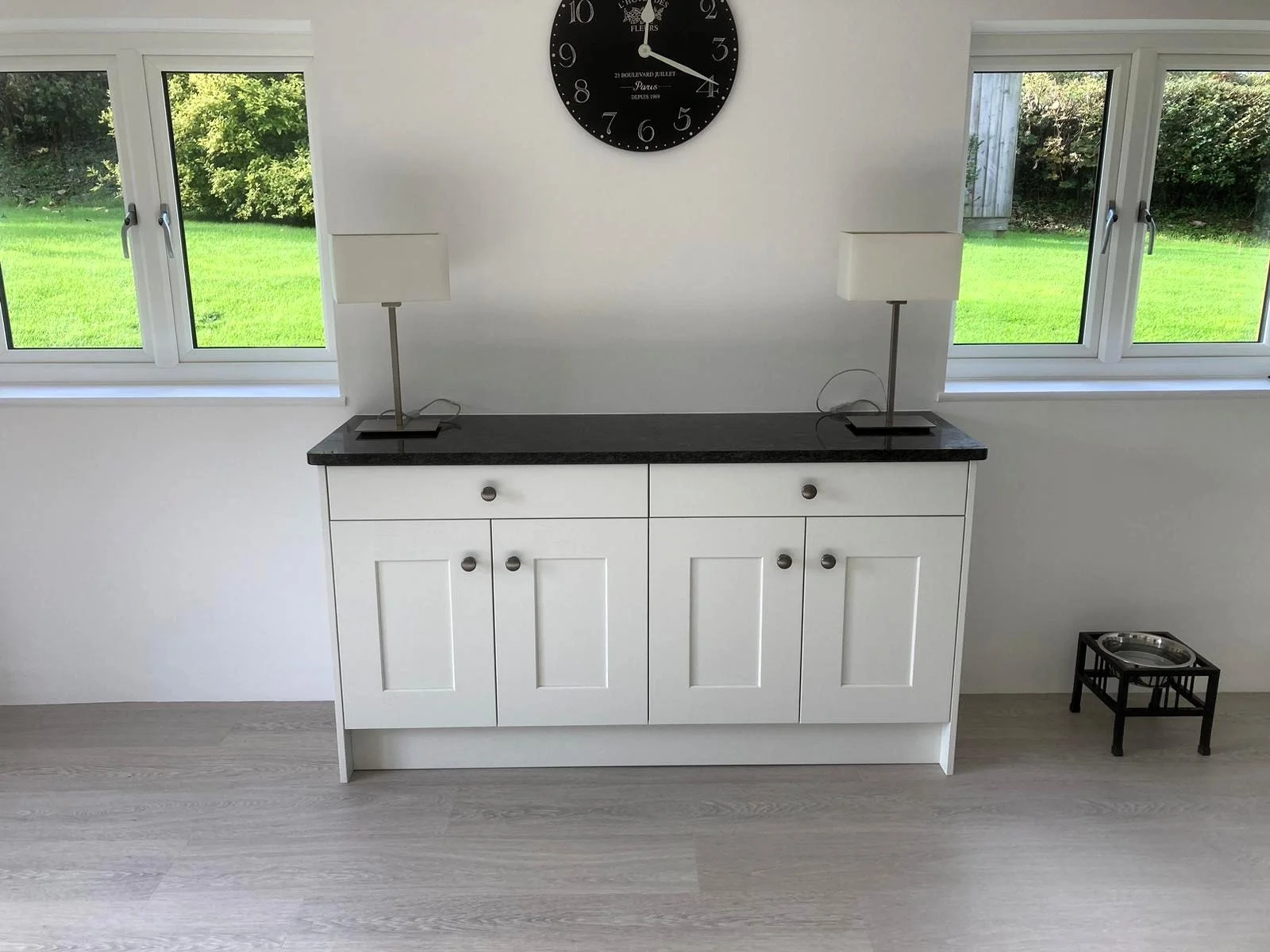 A white cabinet with a black countertop positioned in front of a white wall and two windows showcasing a green outdoor yard. 