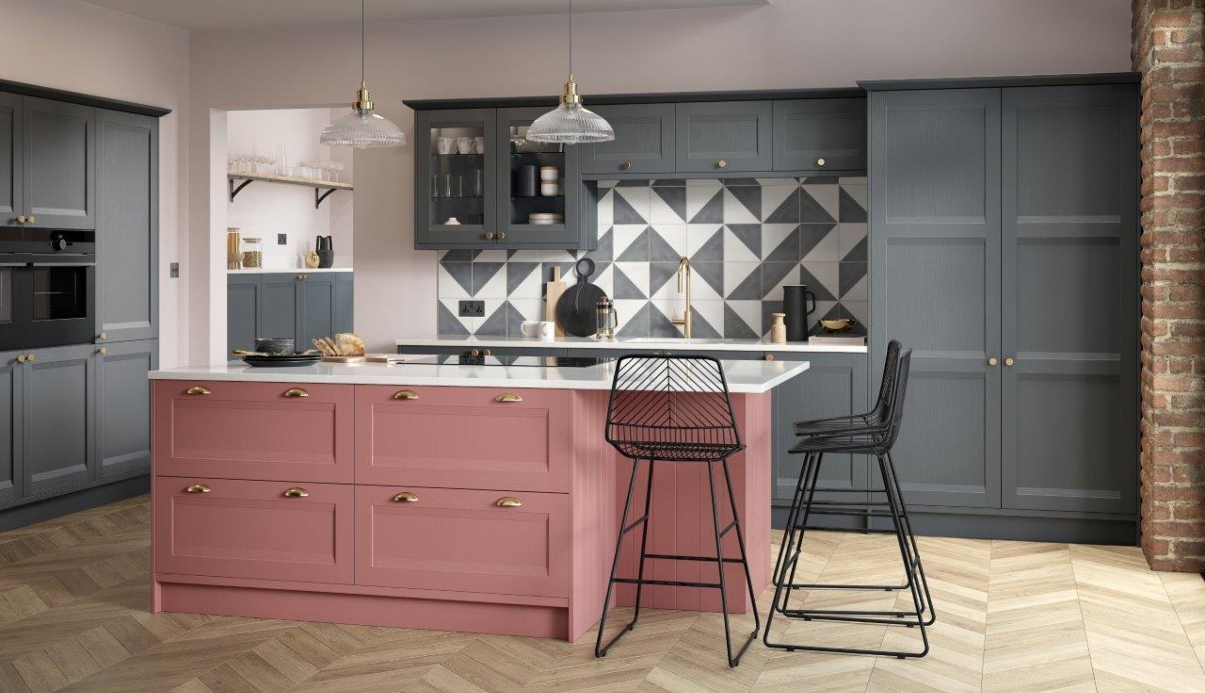 Marpatt kitchen with grey cabinetry, a pink kitchen island with gold handles, black chairs, and a brick wall on the right. Geometric tile backsplash behind the sink, with hanging pendant lights above the island.