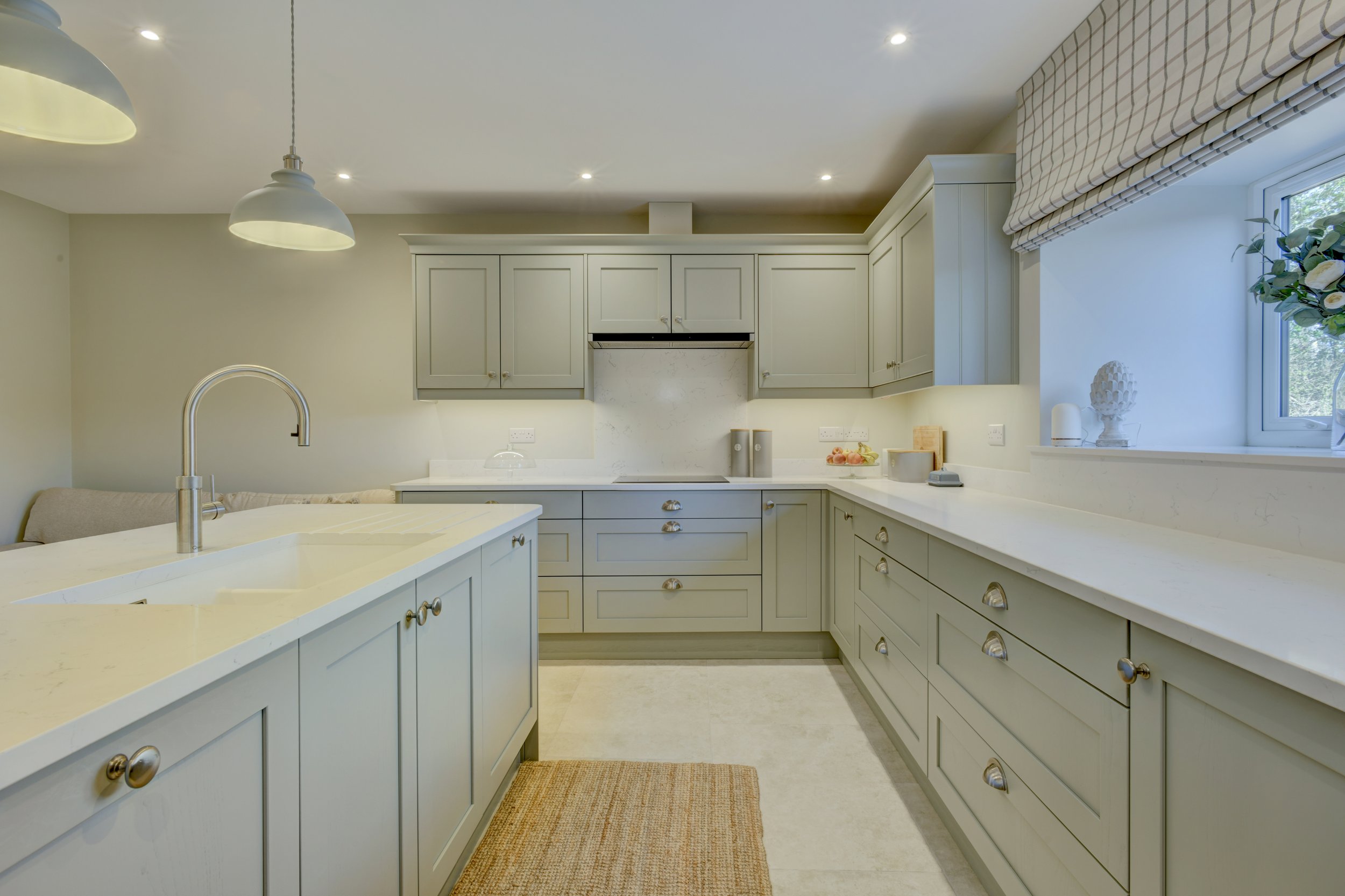 A contemporary kitchen with greige cabinets, Quartz countertops, a large window with a checkered Roman shade, and a beige rug on the floor.