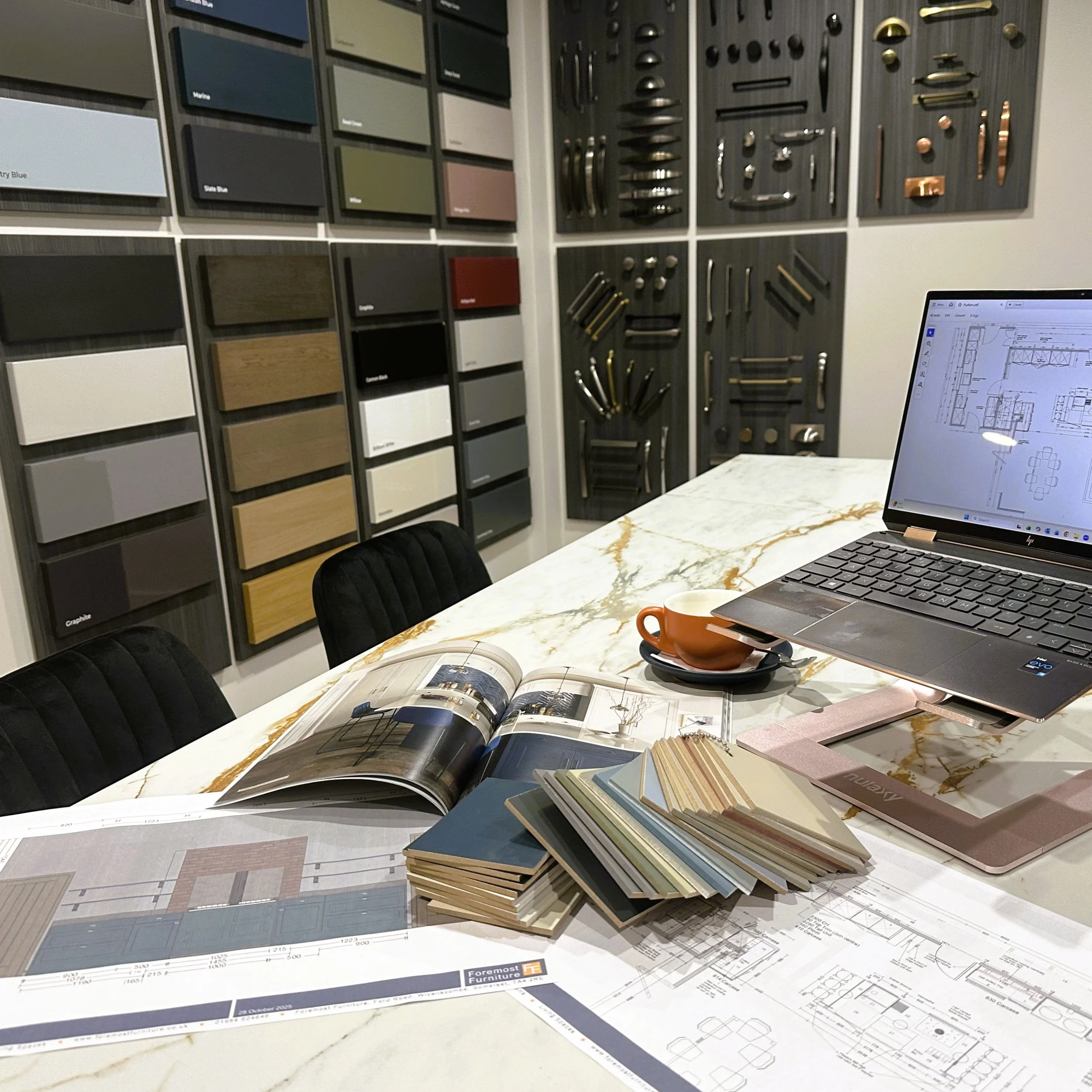 Interior design studio with wall samples of door colors, and handle finishes. A laptop displaying architectural drawings, and an open magazine on a granite table.