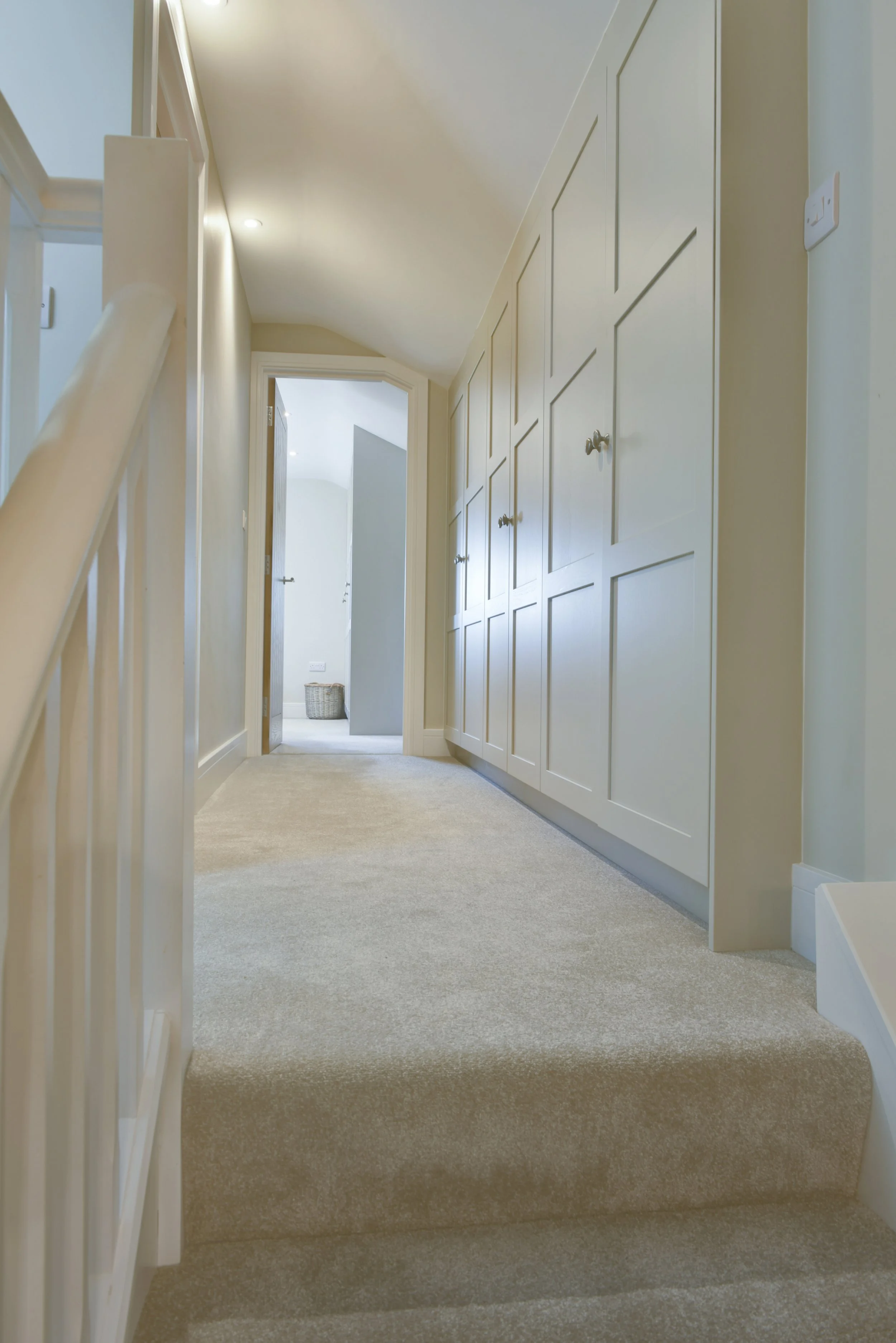 A hallway with cream-colored carpet and built-in cream wardrobes on the right. An open door at the end leads to a sunlit bedroom with a basket on the floor.