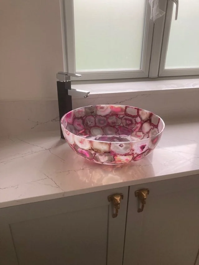 A decorative pink and white marble sink in a bathroom on a white countertop near a window with frosted glass.