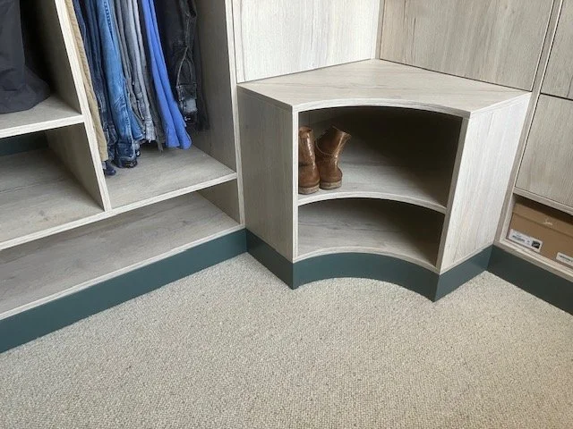 A corner of a walk-in closet with a small shelf containing a pair of brown boots, next to a shoe rack with folded clothes and hanging jeans.