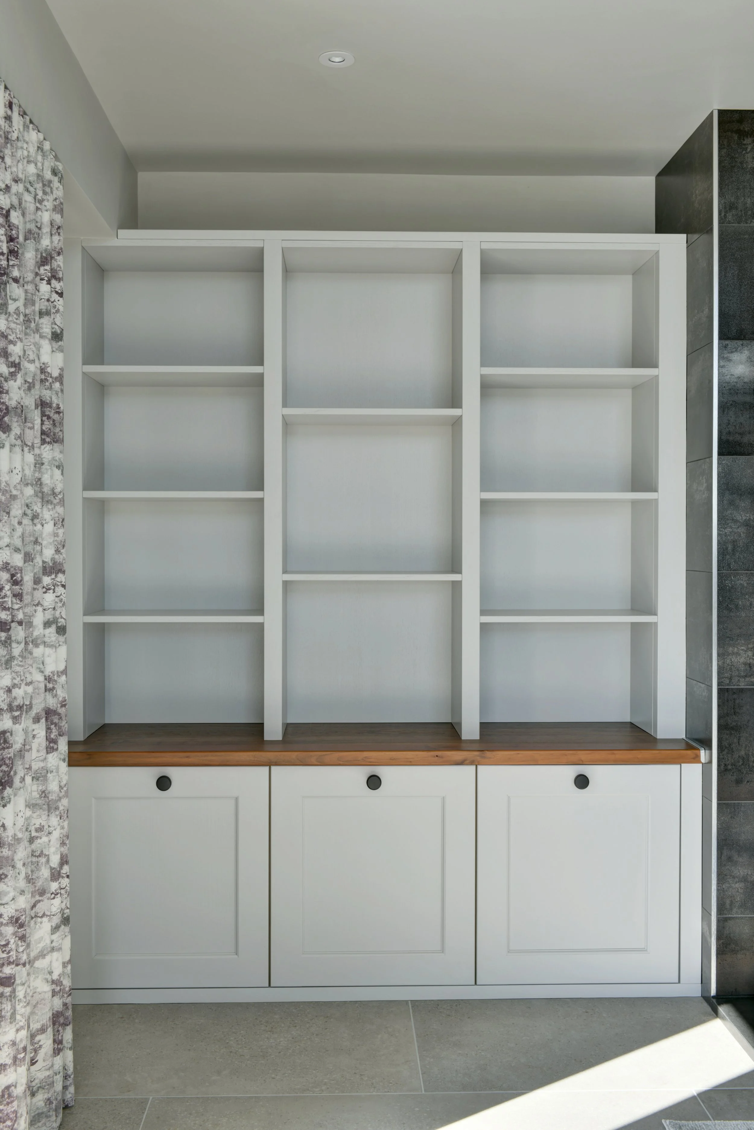 White bookshelf unit with three shelves and three drawers at the bottom, against a wall with patterned curtains on the left and a dark tiled wall to the right.