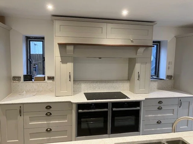 Kitchen with white cabinets and central mantel with two small windows wither side. Built in double oven and hob. 