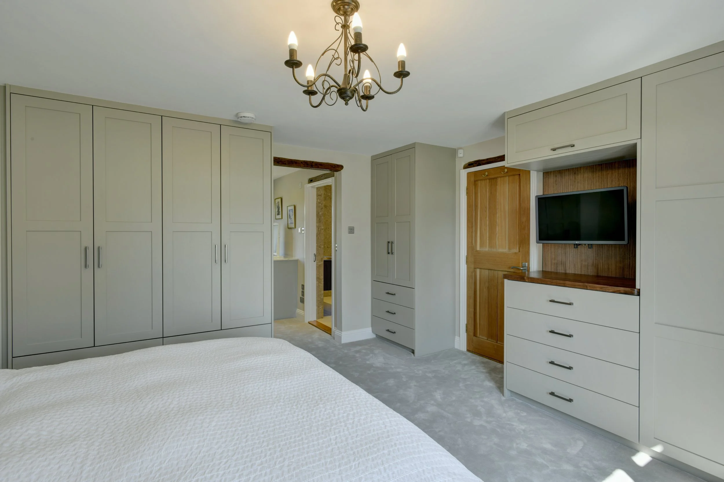 A bedroom with white built-in wardrobes, a wooden door, a wall-mounted TV, and a chandelier hanging from the ceiling.