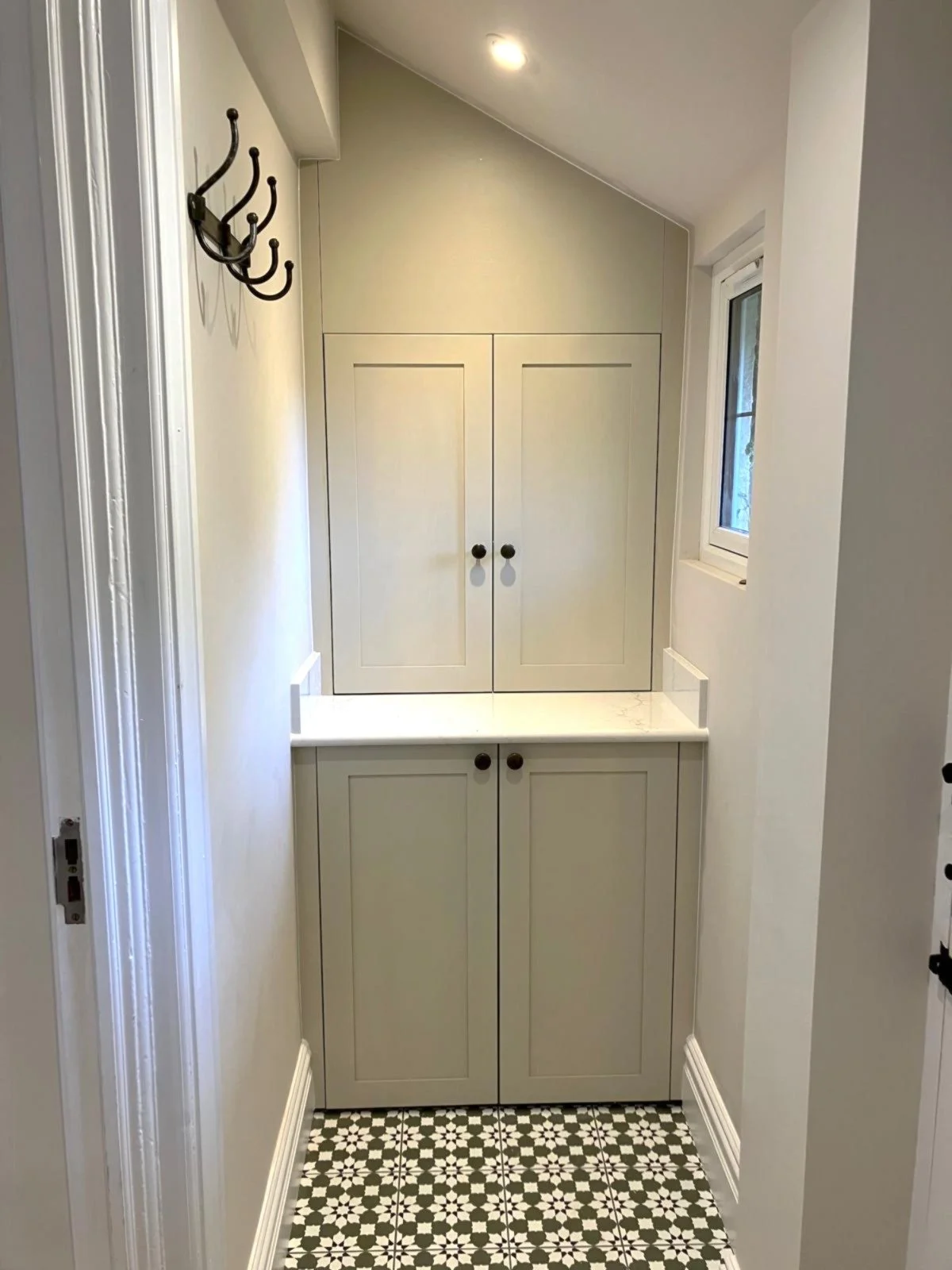 Small utility room with built-in cabinetry, a window, patterned floor tiles, and a coat hook on the wall.