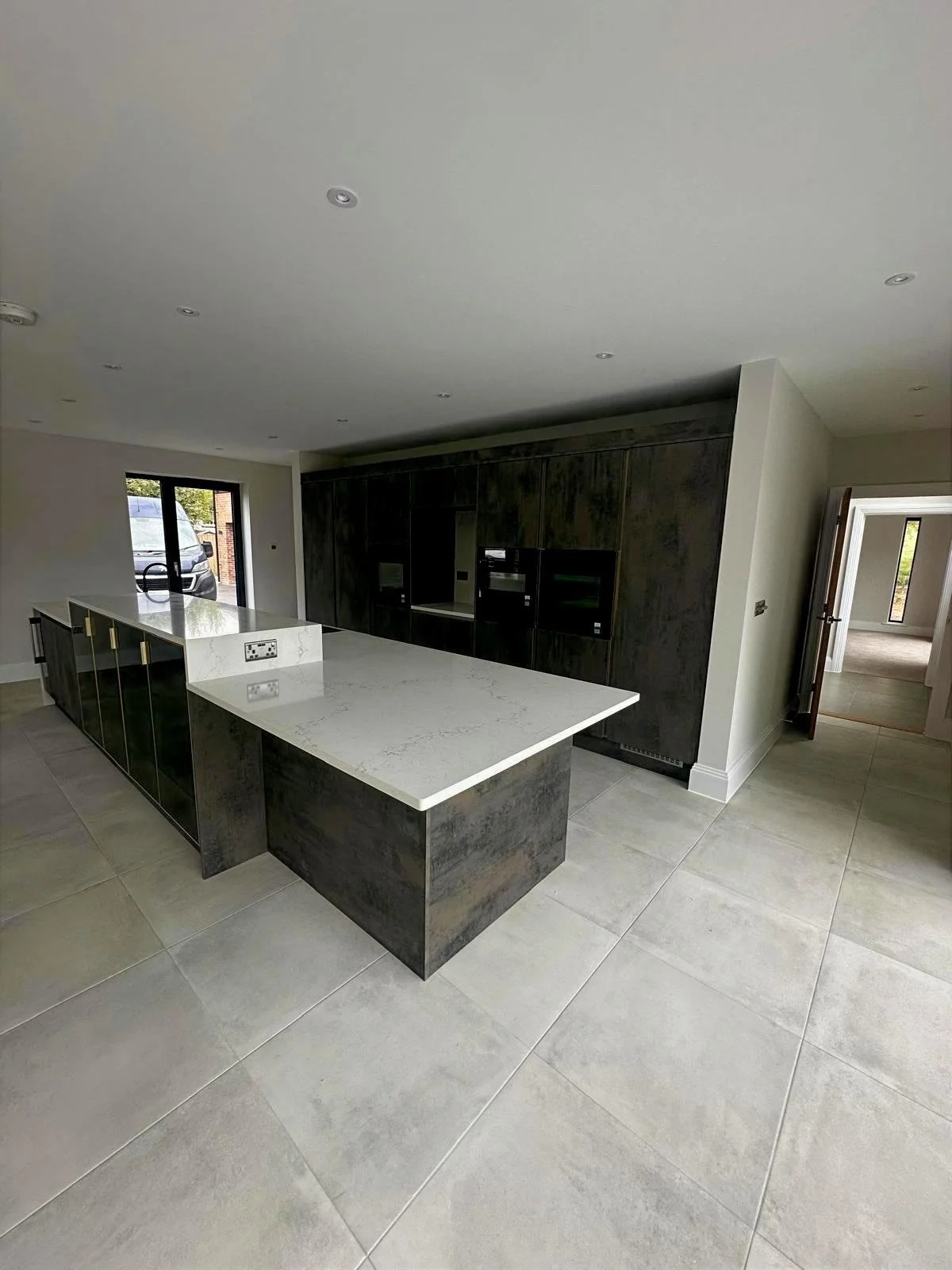 Modern kitchen with large white marble island, dark cabinetry, built-in appliances, tiled floor, and natural light from sliding glass door.