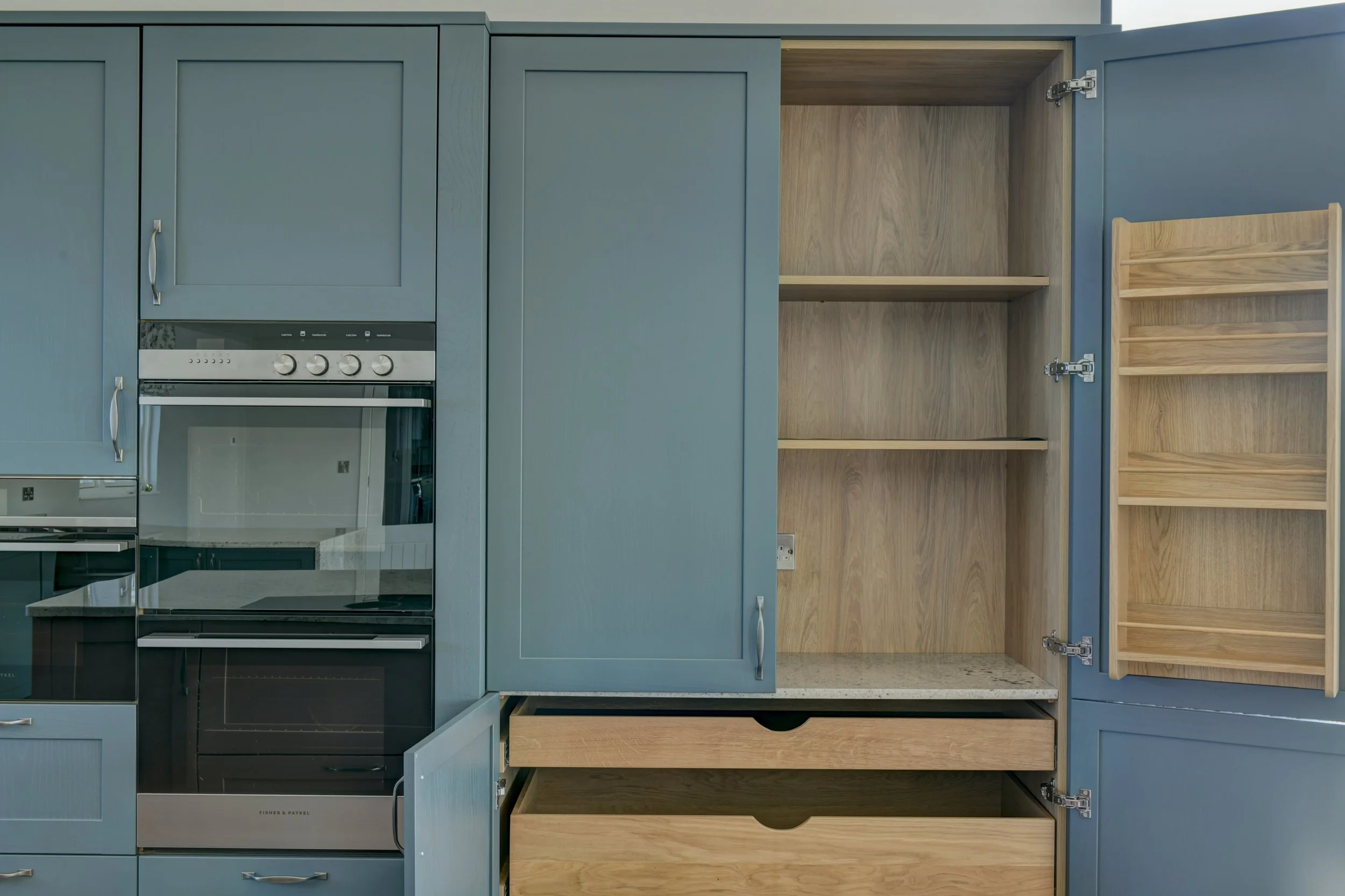 Open blue kitchen cabinet with wooden shelves and drawers inside, adjacent to a built-in oven.