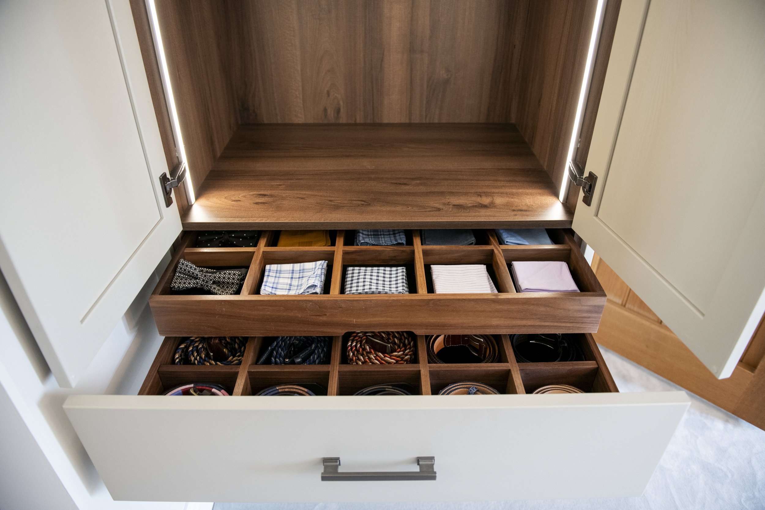 Open white cabinet with wooden interior, empty upper shelf, and organized lower drawer containing folded clothing and accessories.