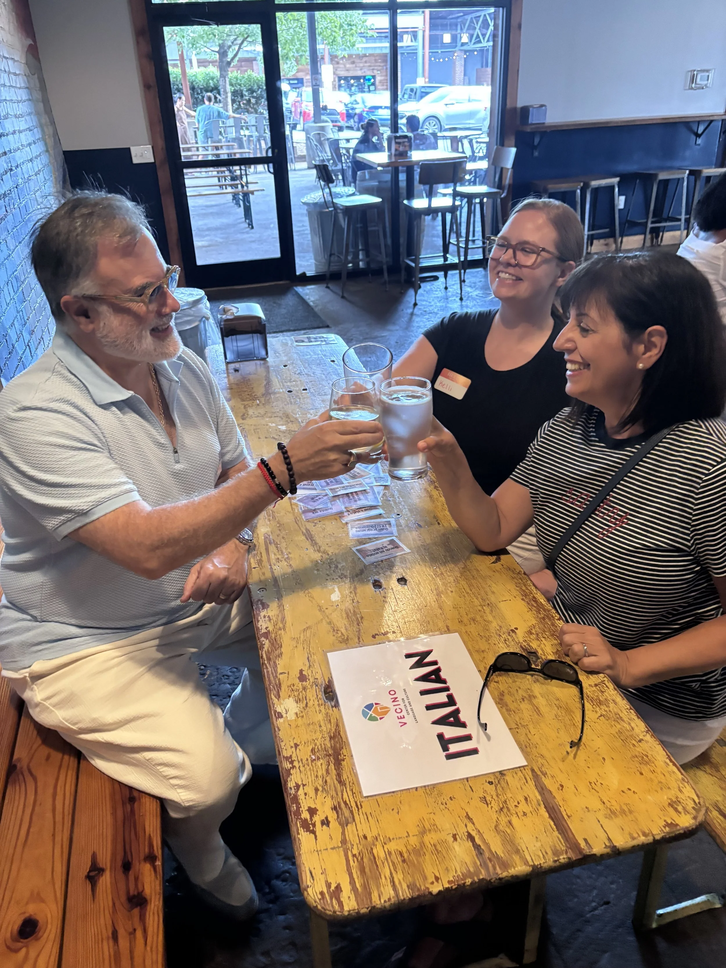 Three Italian speakers sitting at a wooden table in a restaurant, toasting with glasses, smiling and enjoying their time together, with a sign that says 'ITALIAN' on the table.