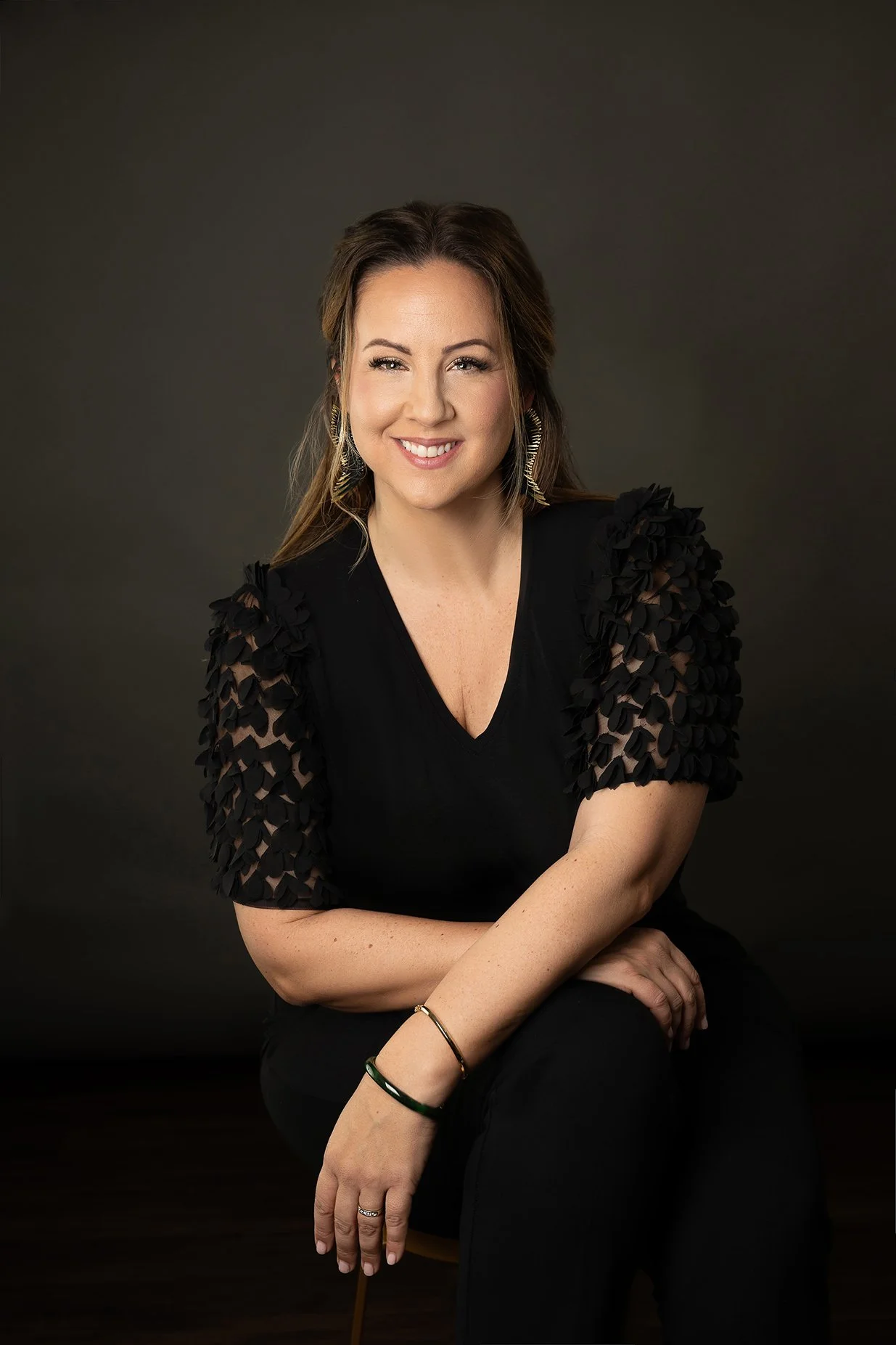 Woman in a black dress sitting against a dark grey backdrop.