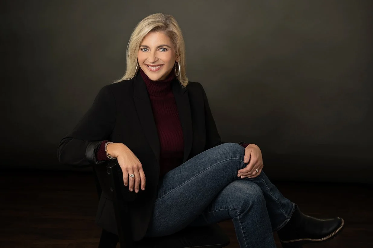 Blonde woman sitting cross-legged in jeans in a chair with a gray background.