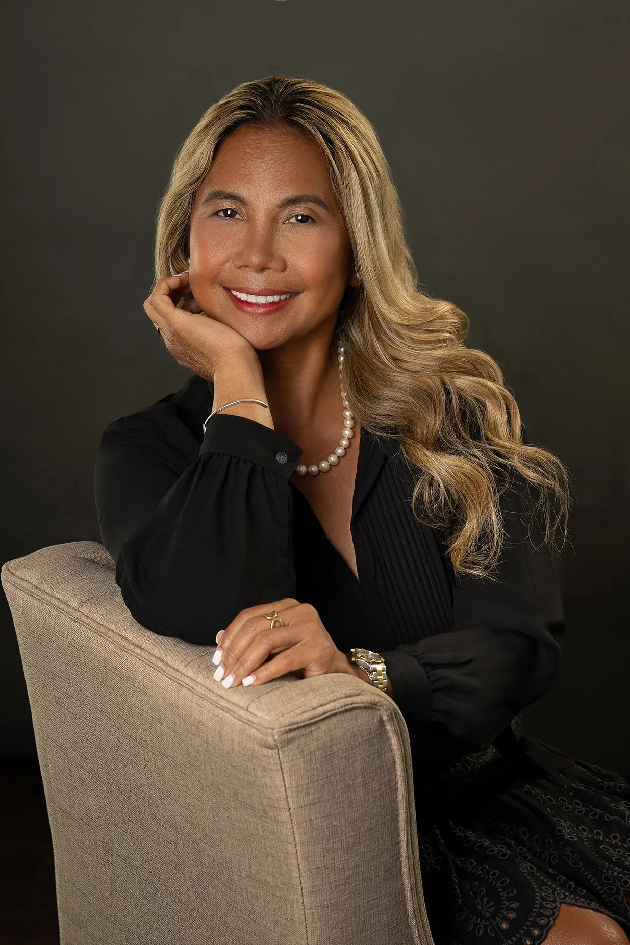Fair haired woman sitting in a brown chair and wearing a black dress with her hand under her chin.