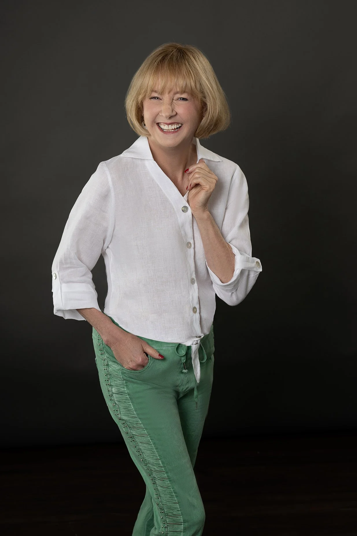 Woman with short blonde hair and a white shirt with green pants against a dark gray background.