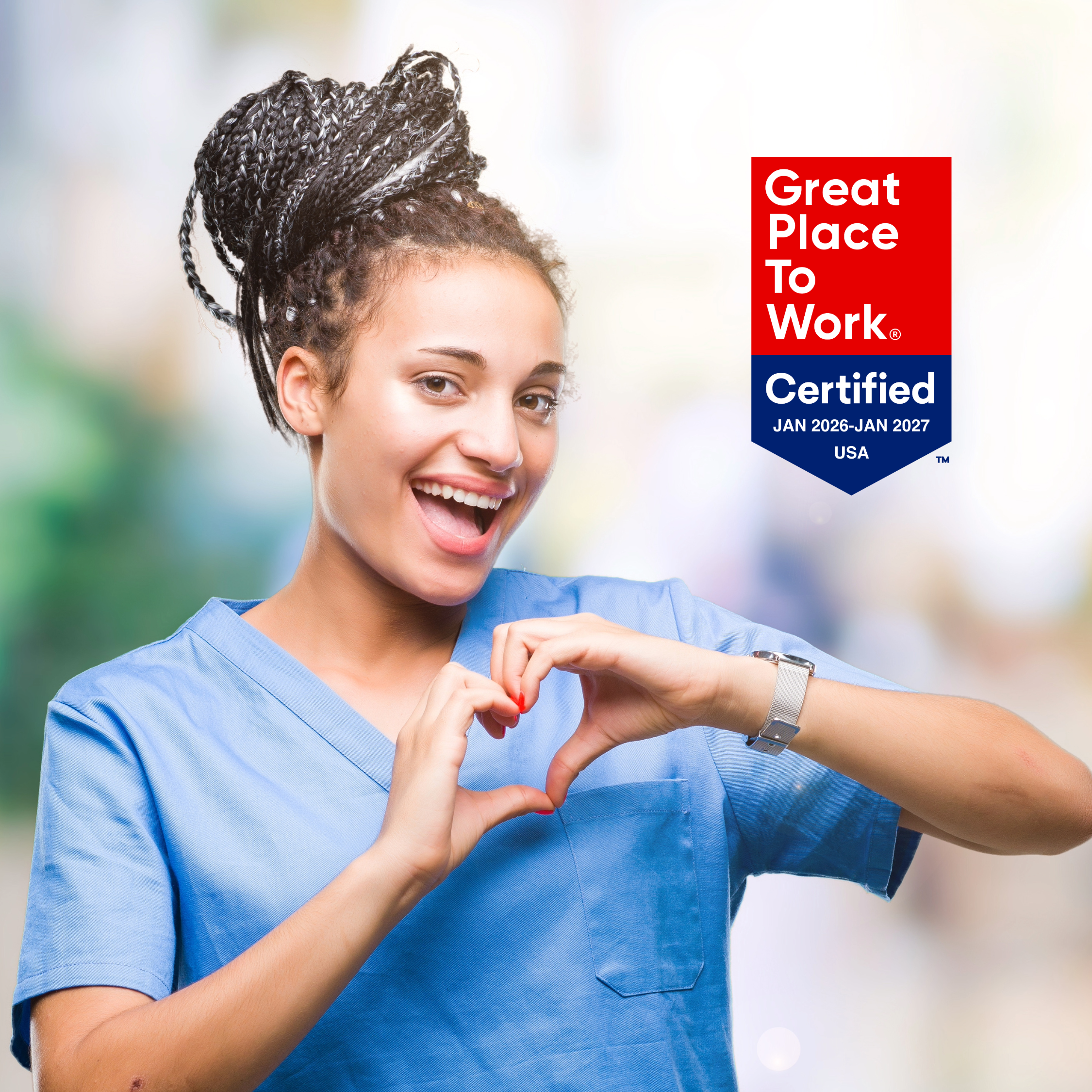 Happy female healthcare worker in blue scrubs making a heart shape with her hands, with a Great Place to Work Certified badge overlapping the top right corner.