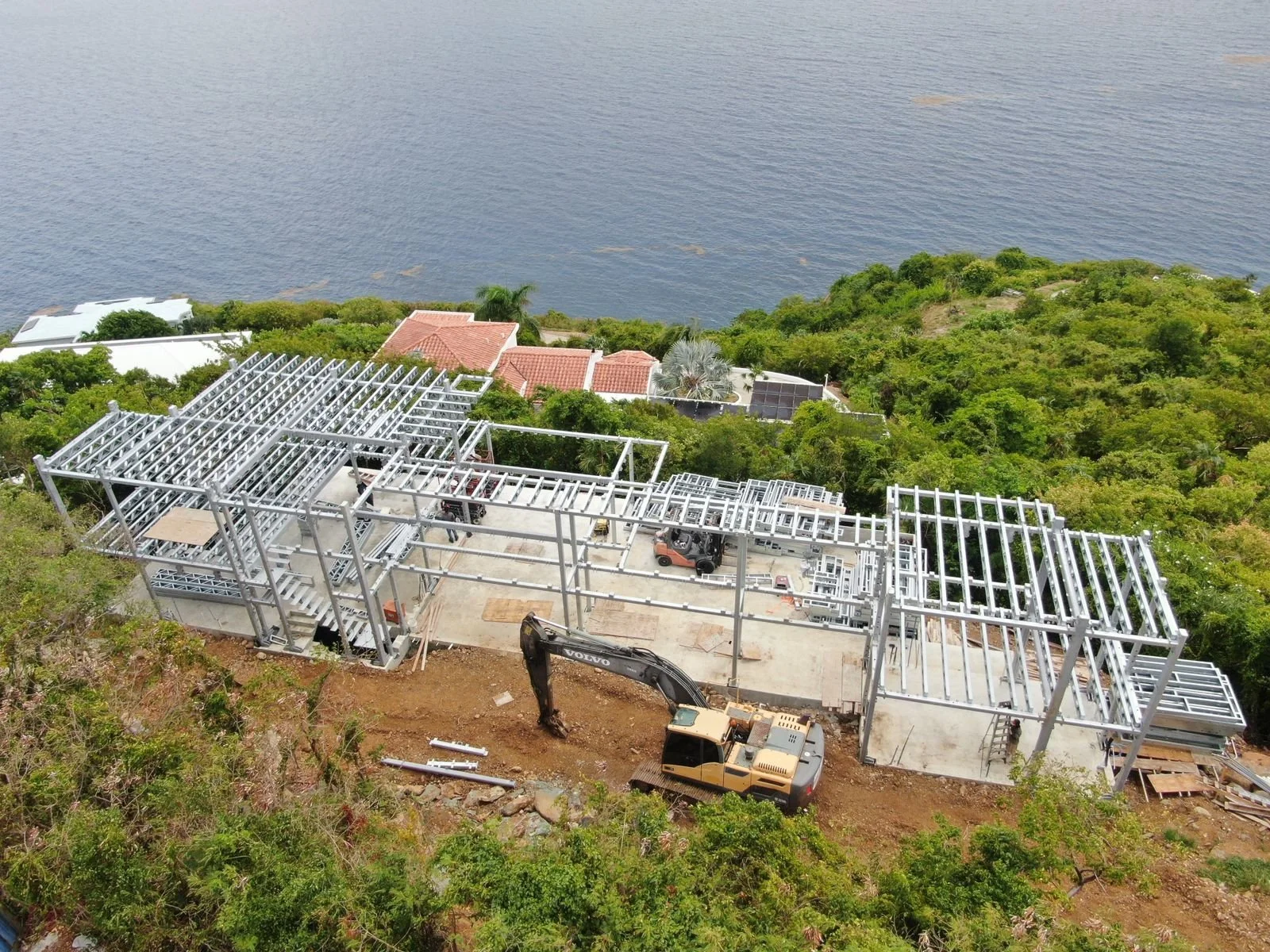 Steel framework for a modular, hurricane resistant steel frame Bauhu house under construction on a hillside near a body of water, with construction equipment and vehicles present.